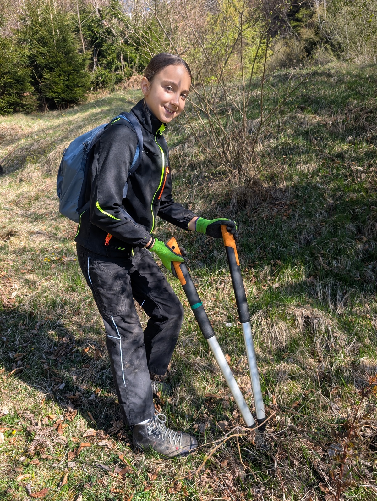 Eine Person in Outdoor-Kleidung und Handschuhen steht auf einer Wiese und hält eine langstielige Baumschere in der Hand. Wahrscheinlich ist sie mit der Almpflege auf der Arzler Alm beschäftigt, mit einem Rucksack und Bäumen im Hintergrund.