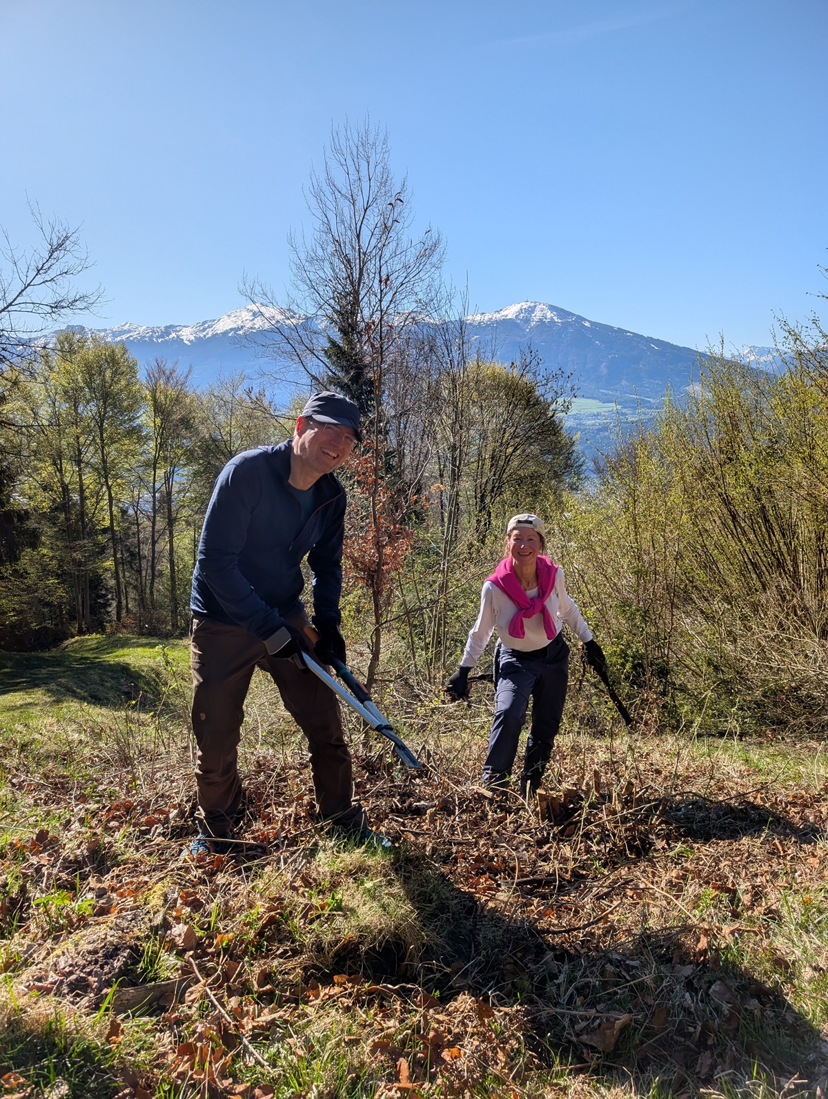 Zwei Personen sind an einem sonnigen Tag auf der Arzler Alm bei der Gartenarbeit im Freien und üben Almpflege mit Bergen und Bäumen im Hintergrund. Der Mann hält eine Schaufel und die Frau hält Gartengeräte.