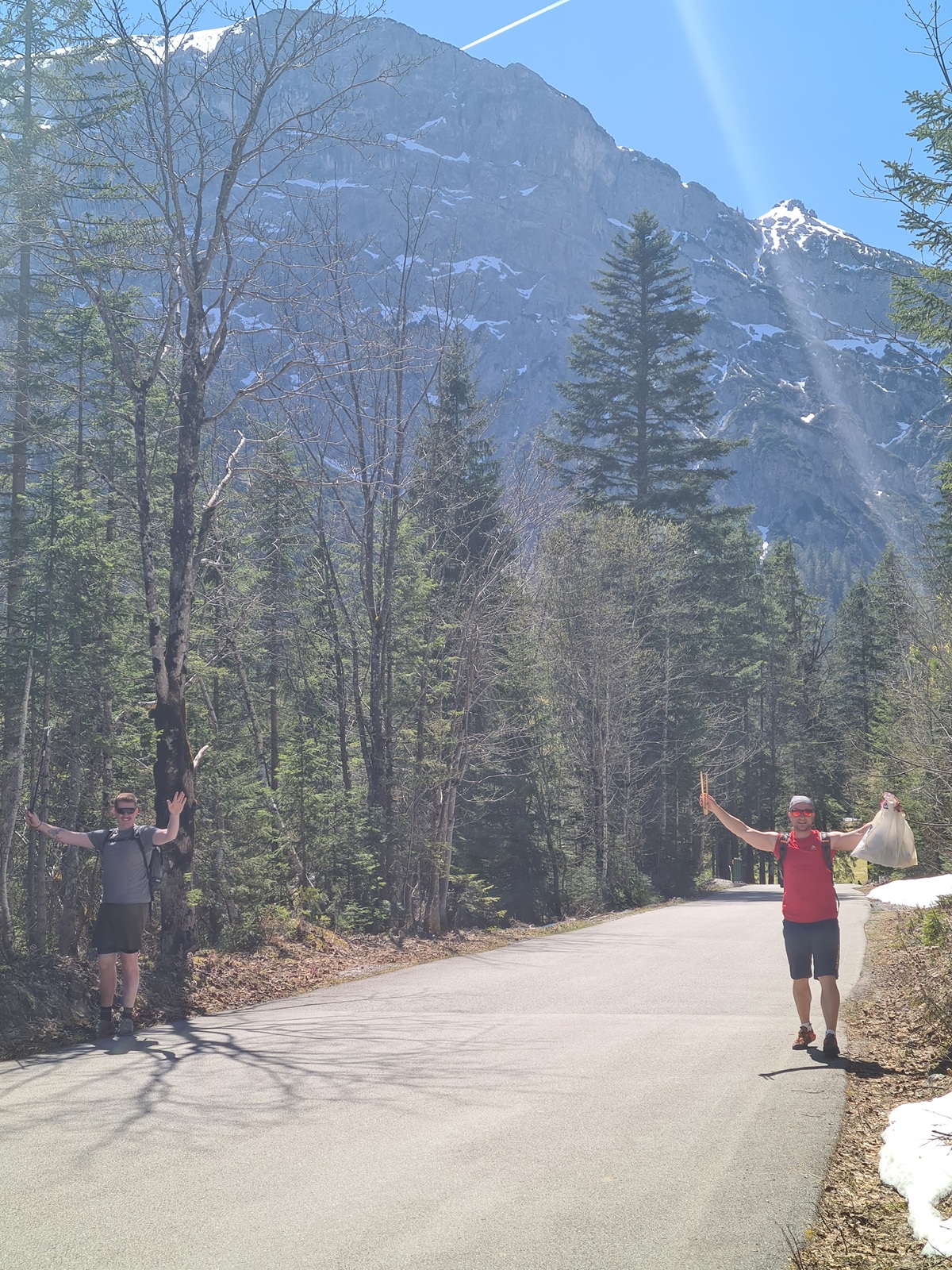 Zwei Personen stehen auf beiden Seiten eines befestigten Weges in den bewaldeten Karwendeltälern in der Nähe des Achensees, mit Schnee auf dem Boden und einem großen felsigen Gipfel im Hintergrund, möglicherweise bei einer Müllsammelaktion.