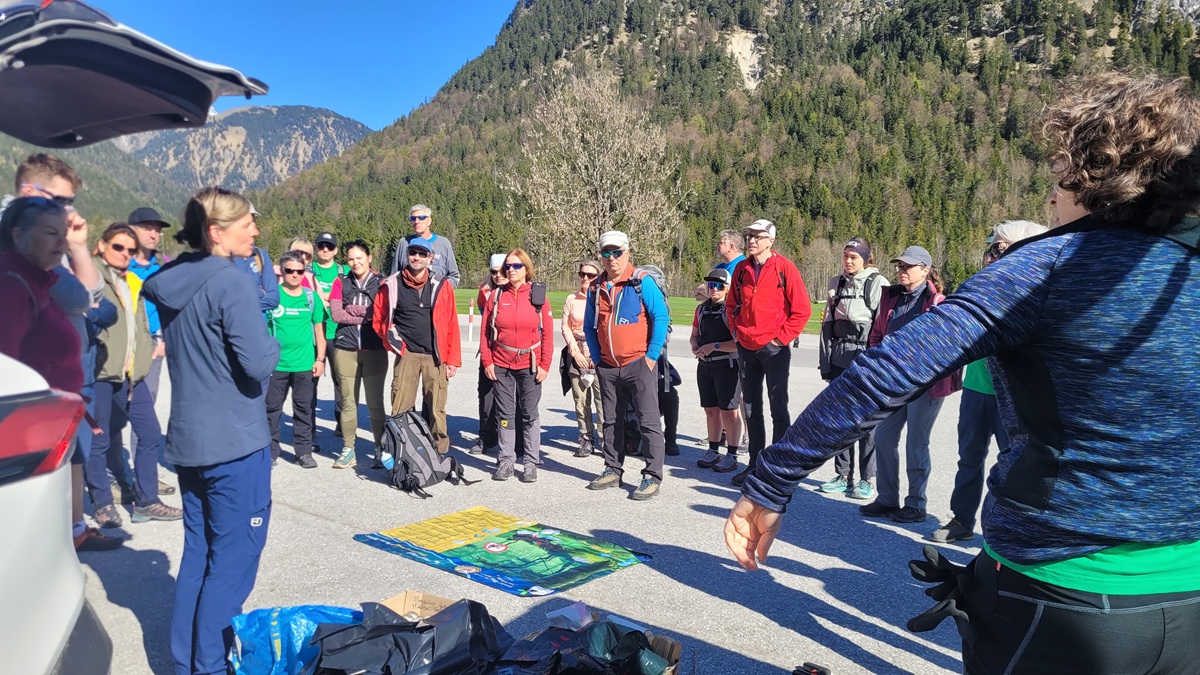 Eine Gruppe von Wanderern mit Rucksäcken lauscht an einem sonnigen Tag auf einem Parkplatz im Freien, umgeben von Bergen und Bäumen, einem Führer und bereitet sich auf eine Müllsammelaktion in den Karwendeltälern am Achensee vor.