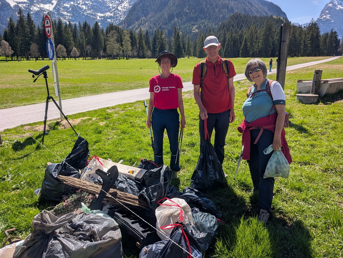 Drei Personen stehen im Freien auf einer Wiese neben gefüllten Müllsäcken und gesammeltem Abfall nach einer Müllsammelaktion im Karwendeltäler Achenseegebiet, im Hintergrund sind Berge, Bäume und ein Parkverbotsschild zu sehen.