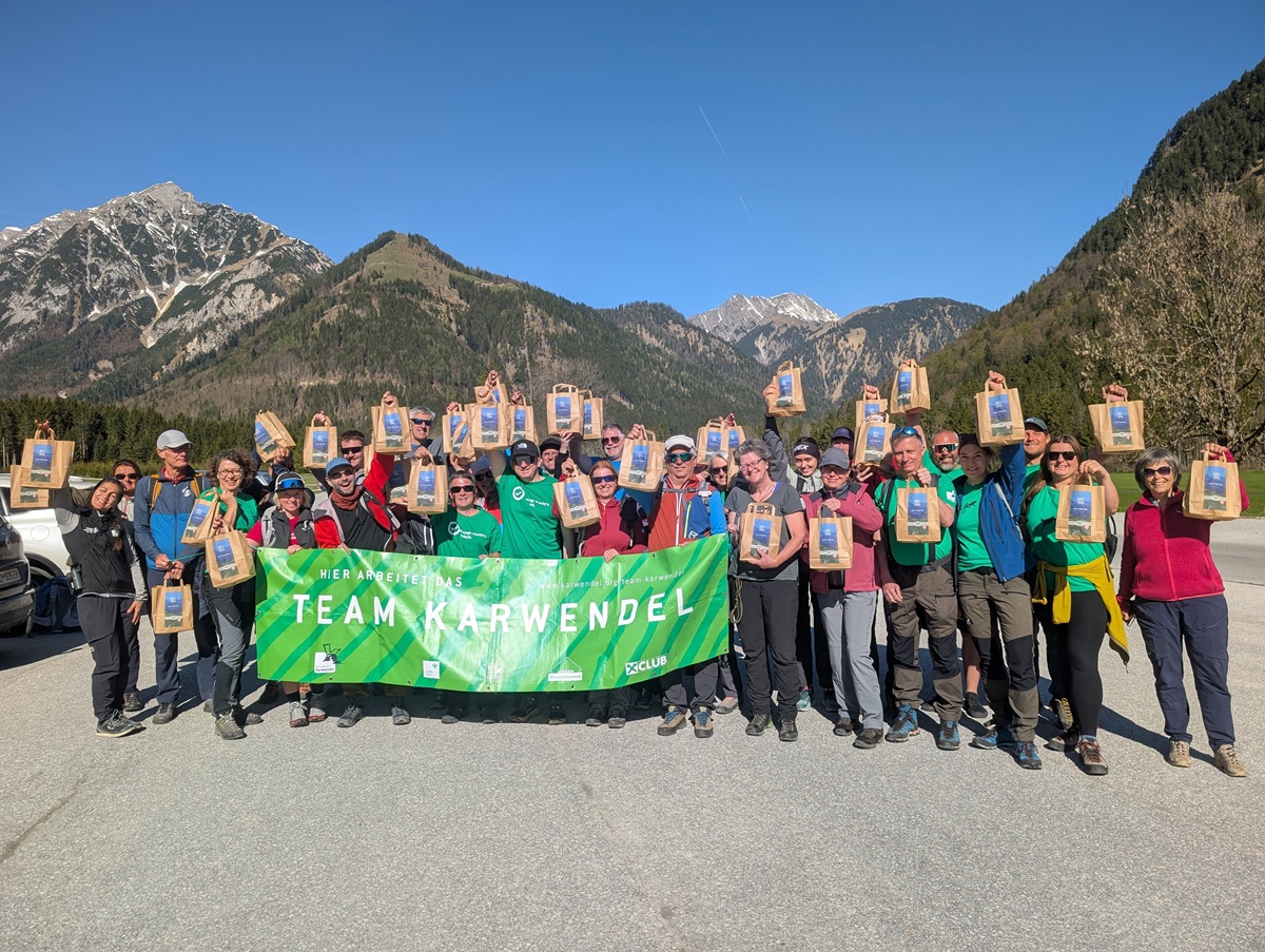 Eine Gruppe von Wanderern steht im Freien vor den Bergen und hält Plaketten und ein grünes Banner mit der Aufschrift "Team Karwendel" nach einer Müllsammelaktion Karwendeltäler Achensee.