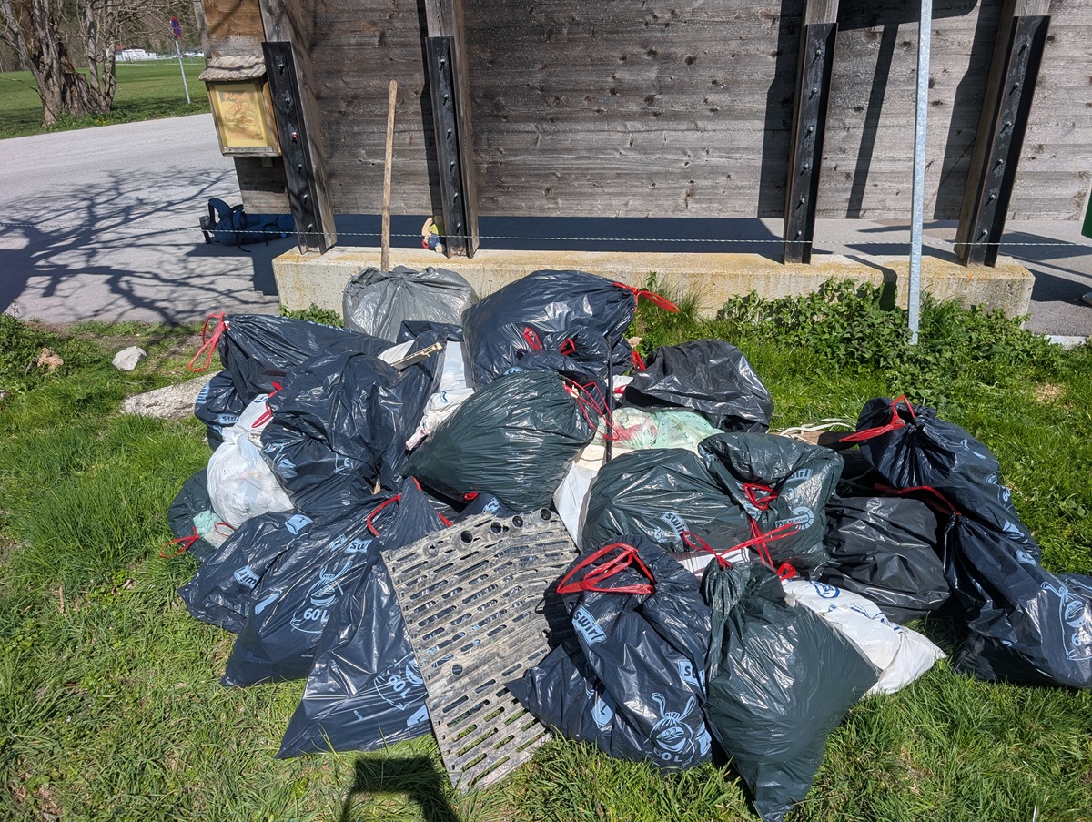 Ein Haufen gefüllter schwarzer Müllsäcke und einige weiße Säcke liegen auf einer Wiese in der Nähe einer Holzkonstruktion, in der Nähe befinden sich ein Metallgitter und Werkzeuge - Beweise für eine kürzlich durchgeführte Müllsammelaktion der Karwendeltäler Achensee.