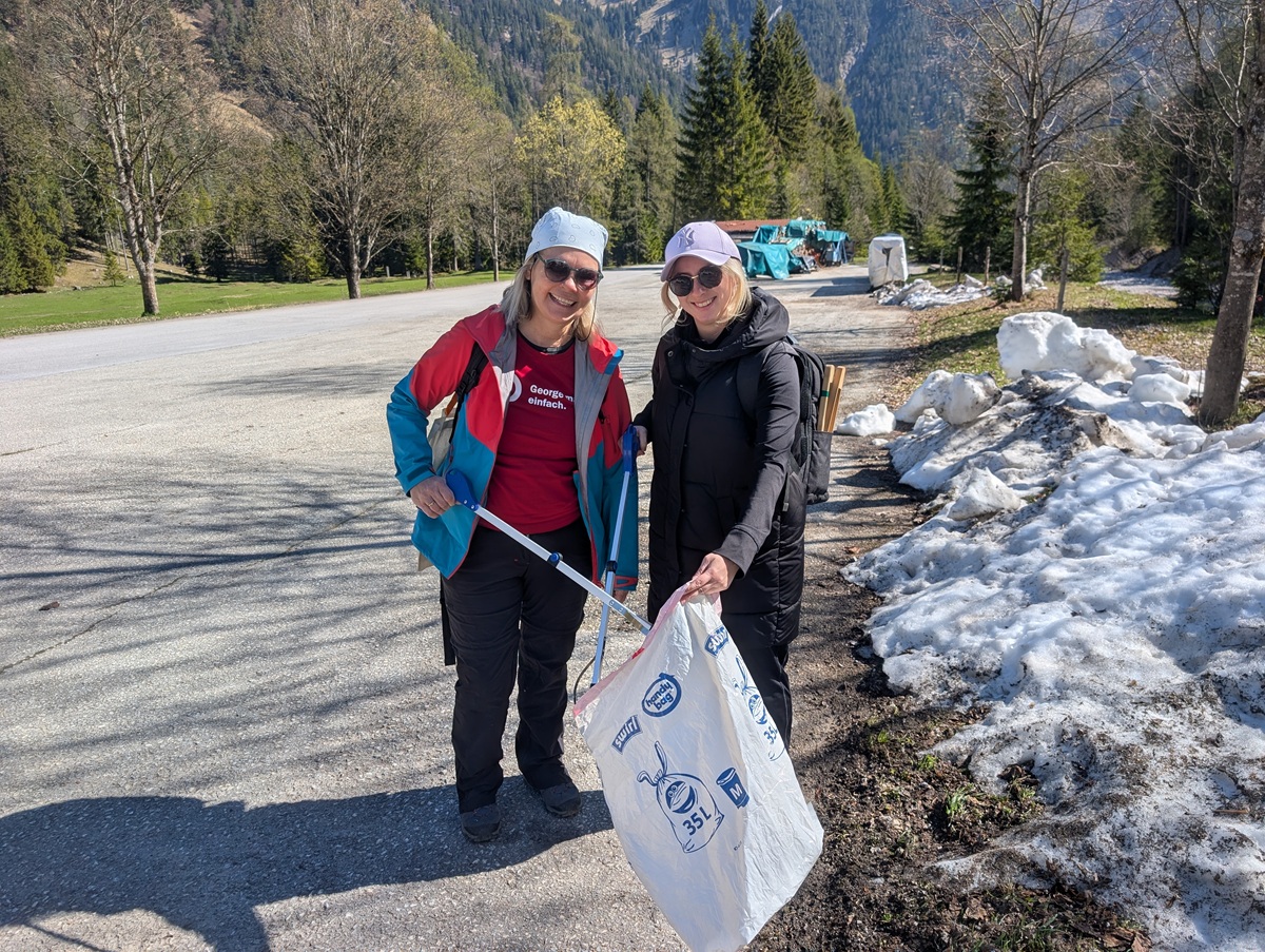 Zwei Frauen in Outdoor-Kleidung sammeln bei der Müllsammelaktion Karwendeltäler Achensee mit Greifer und Sack am verschneiten Straßenrand Müll ein, im Hintergrund Bäume und Berge.