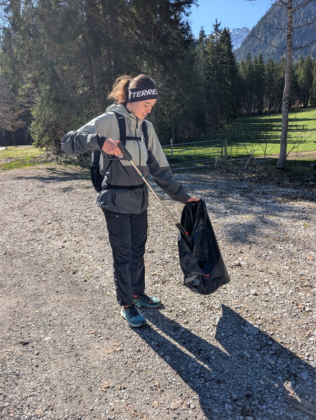 Bei einer Müllsammelaktion in den Karwendeltälern in der Nähe des Achensees sammelt eine Person in Outdoor-Kleidung und mit Stirnband auf einem Schotterweg mit Bäumen und Bergen im Hintergrund mit einer Greifzange Müll auf und legt ihn in einen schwarzen Sack.