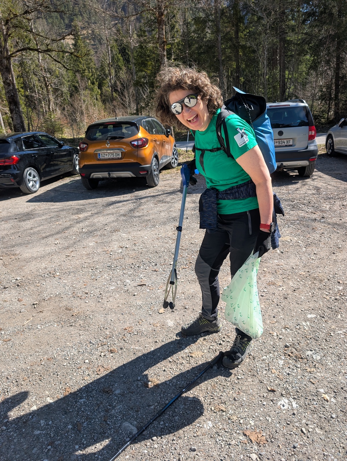 Person in Outdoor-Kleidung, Sonnenbrille und Rucksack steht mit Trekkingstöcken und grüner Plastiktüte auf einem Schotterparkplatz - bereit für die Müllsammelaktion Karwendeltäler Achensee; im Hintergrund sind Autos und Bäume zu sehen.