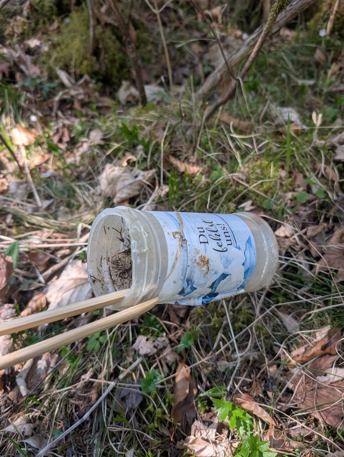 Ein weggeworfener Plastikbehälter mit blau-weißer Aufschrift liegt inmitten von Gras, Laub und Stöcken in der freien Natur auf dem Boden und weist auf die Notwendigkeit einer Müllsammelaktion Karwendeltäler Achensee hin, um die Natur sauber zu halten.