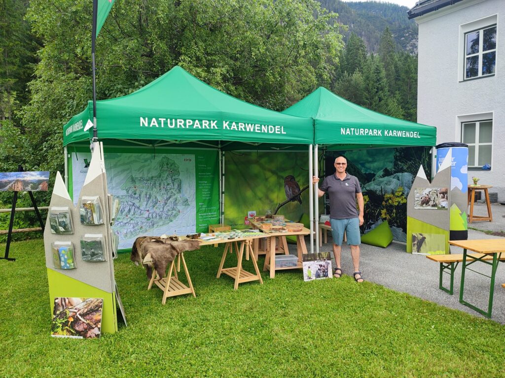 Ein Mann steht neben einem Informationsstand des Naturparks Karwendel mit grünen Zelten, Schautafeln, Broschüren und Karten, die im Freien auf einer Wiese aufgestellt sind.