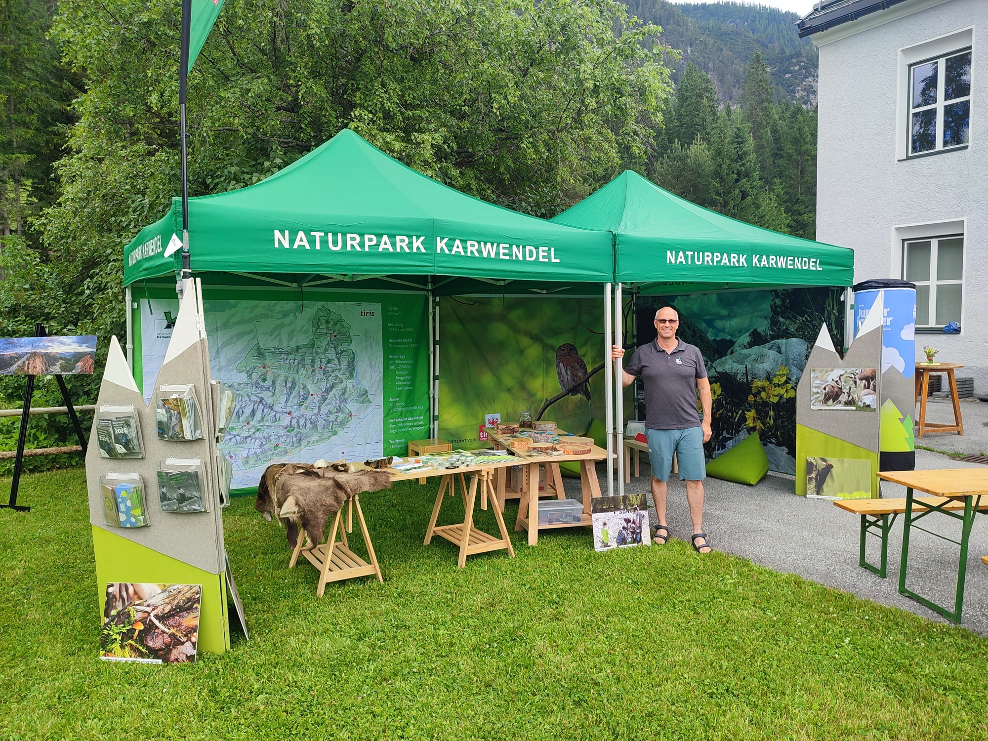 Ein Mann steht neben einem Informationsstand des Naturparks Karwendel mit grünen Zelten, Schautafeln, Broschüren und Karten, die im Freien auf einer Wiese aufgestellt sind.