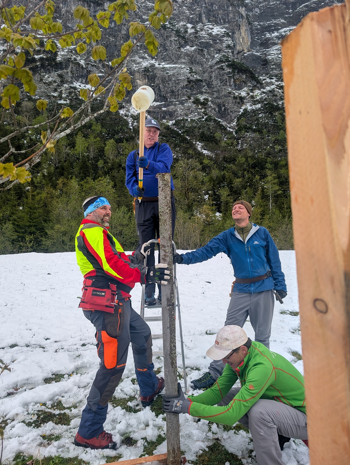 Vier Personen installieren im Freien in verschneitem Gelände Ausrüstung an einem Holzpfosten; einer hält eine lange Stange mit einem Gerät an der Spitze, während die anderen den Pfosten sichern, wahrscheinlich als Hilfe bei Zaunreparaturen am Ahornboden.