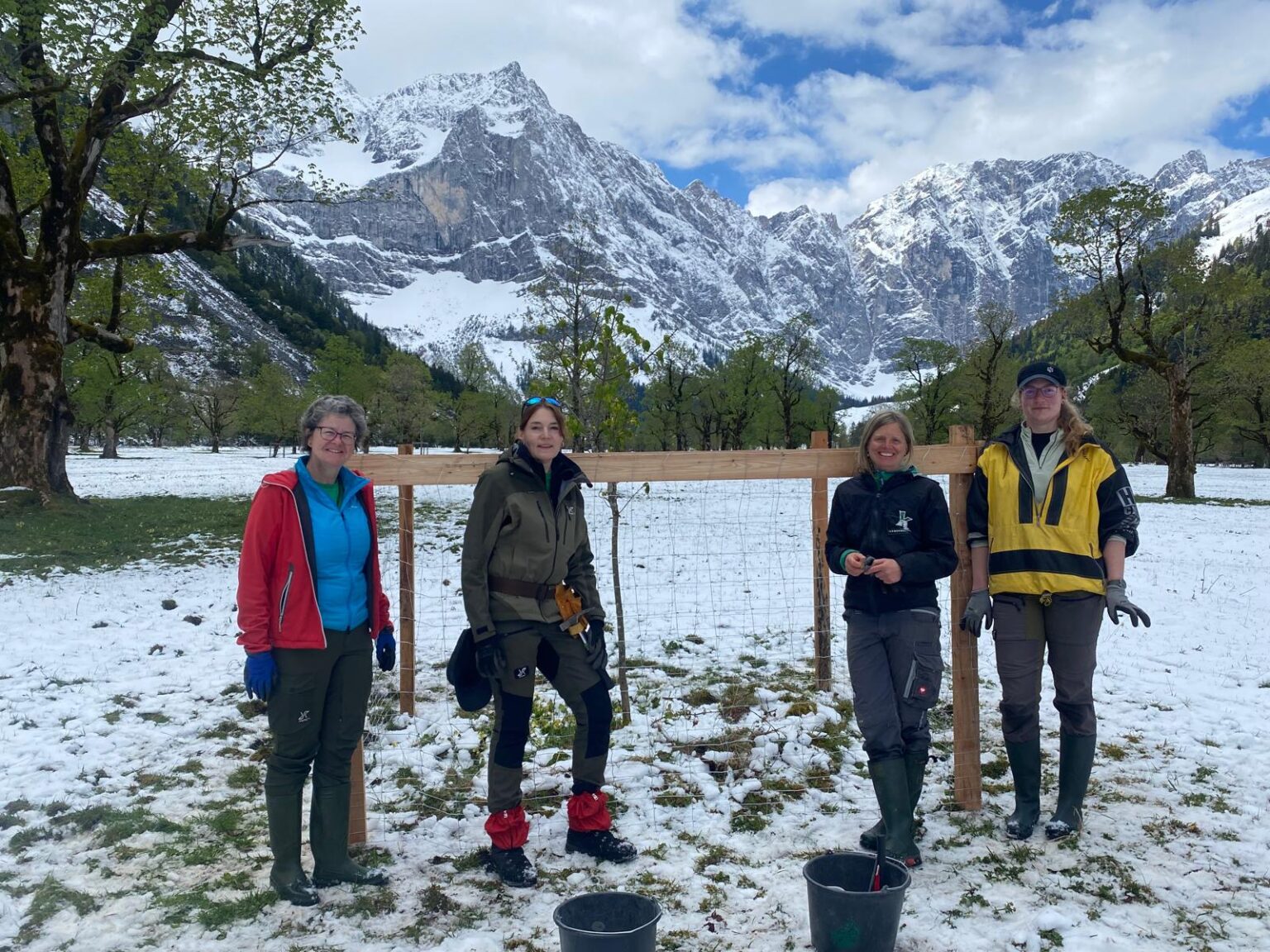 Vier Personen in Outdoor-Ausrüstung stehen vor einem Holzzaun, möglicherweise inmitten der Zaunreparaturen Ahornboden, umgeben von verschneiten Bergen, Bäumen und Wolken im Hintergrund.
