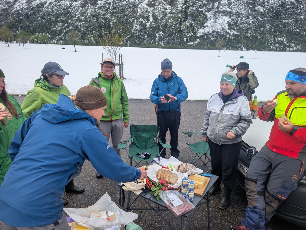 Eine Gruppe von Menschen in Outdoor-Ausrüstung genießt Sandwiches und Snacks an einem Klapptisch, der neben geparkten Autos im verschneiten, bergigen Ahornbodengebiet aufgestellt ist, und macht möglicherweise eine Pause von Zaunreparaturen.