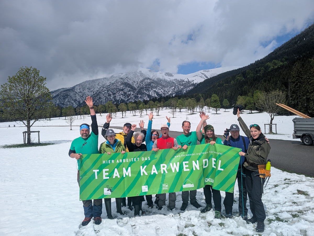 Eine Gruppe von Menschen in Winterkleidung steht im Schnee und hält ein grünes "Team Karwendel"-Banner in die Höhe. Im Hintergrund sind verschneite Berge und Bäume zu sehen, die ihr Projekt "Zaunreparaturen Ahornboden" feiern.