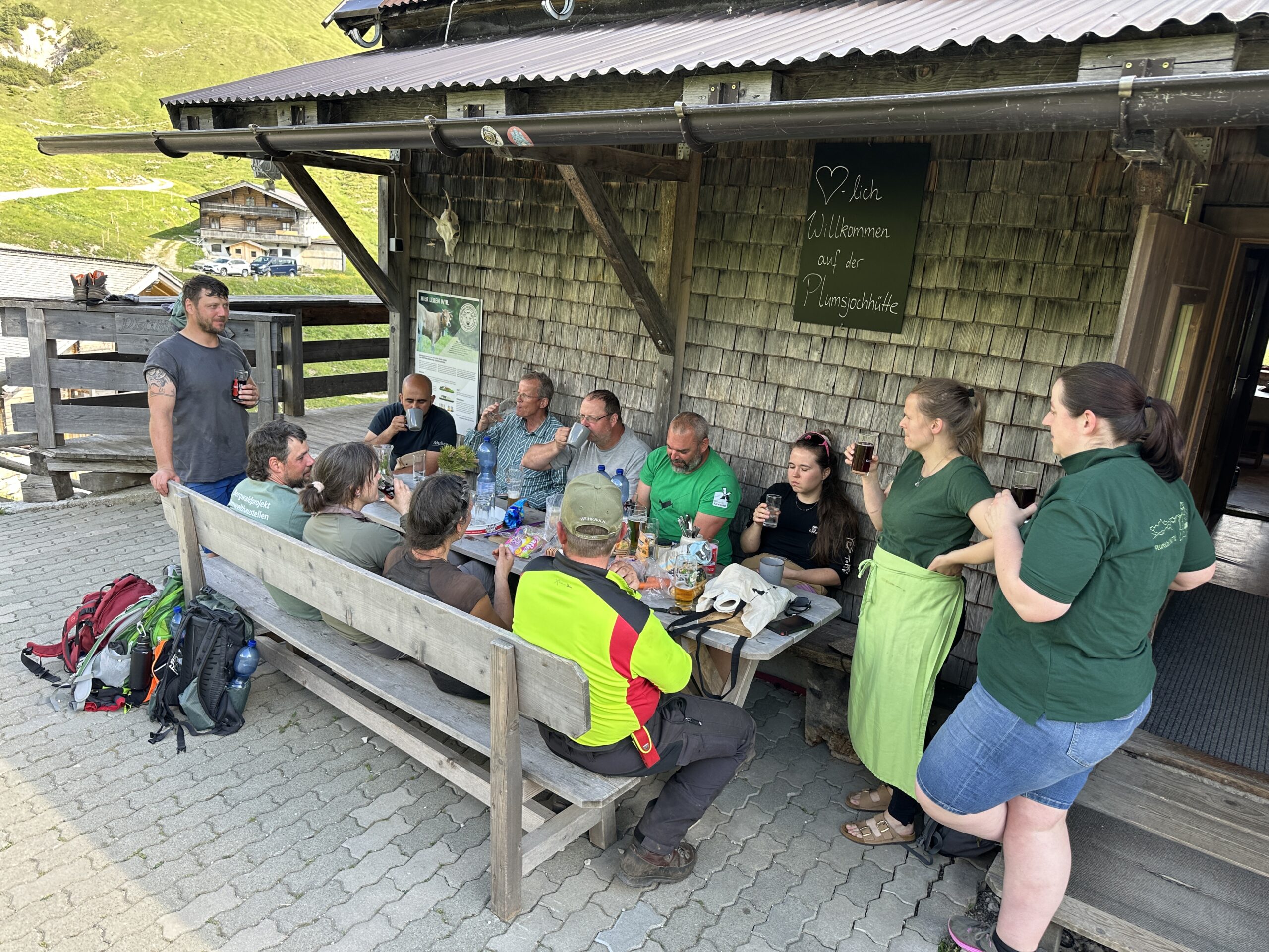 Bergwaldprojekt Umweltbaustelle Almpflege Plumsjochhütte 2026 Team Karwendel Eine Gruppe von Menschen sitzt im Freien um einen Holztisch herum, isst und unterhält sich in der Nähe eines rustikalen Gebäudes mit einer Kreidetafel und Rucksäcken auf dem Boden – möglicherweise ruhen sie sich nach den Zaunreparaturen am Ahornboden aus.