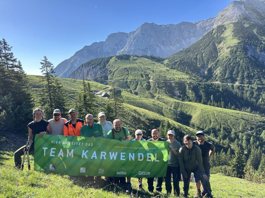 Eine Gruppe von Menschen steht auf einem grasbewachsenen Berghang in der Nähe von Ahornboden, hält ein grünes „Team Karwendel“-Transparent hoch, im Hintergrund sind bewaldete Hügel und felsige Gipfel zu sehen, und feiert die erfolgreichen Zaunreparaturen.