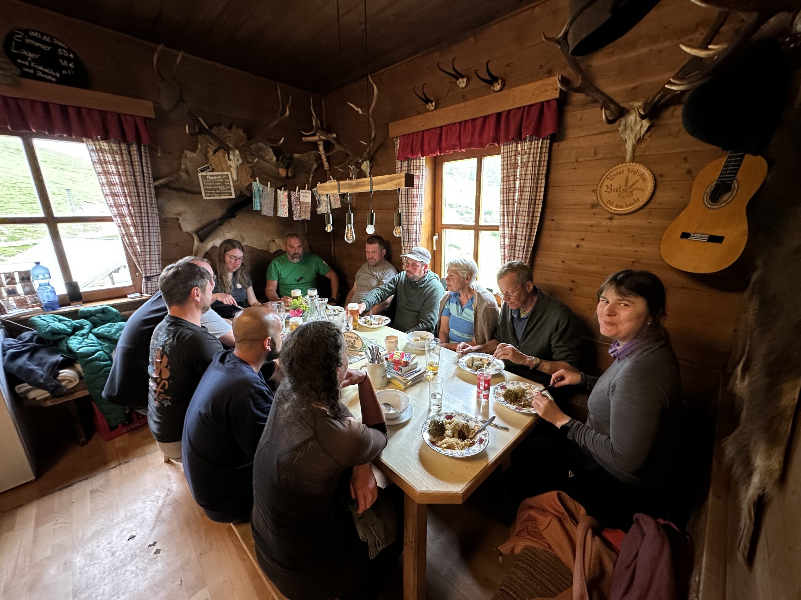 Bergwaldprojekt Umweltbaustelle Almpflege Plumsjochhütte 2026 Team Karwendel Eine Gruppe von Menschen sitzt in einer rustikalen Hütte in der Nähe von Zaunreparaturen Ahornboden um einen Holztisch herum und nimmt gemeinsam eine Mahlzeit ein. An der Wand hängen Geweihe, eine Gitarre und karierte Vorhänge schmücken das gemütliche Interieur.