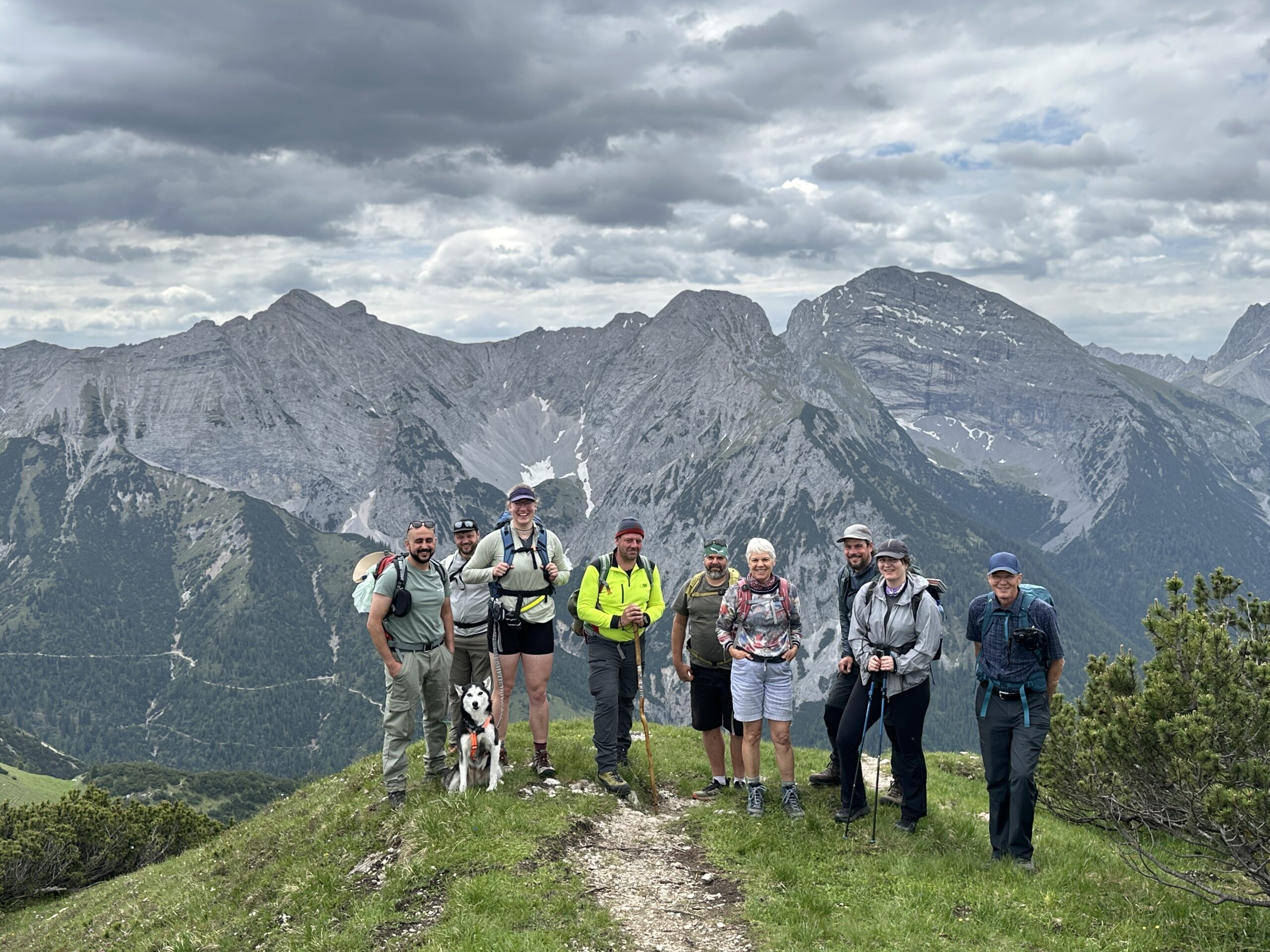 Bergwaldprojekt Umweltbaustelle Almpflege Plumsjochhütte 2026 Team Karwendel Eine Gruppe von acht Wanderern und einem Hund steht auf einem grasbewachsenen Bergweg in der Nähe von Zaunreparaturen Ahornboden, im Hintergrund sind felsige Gipfel und bewölkter Himmel zu sehen.