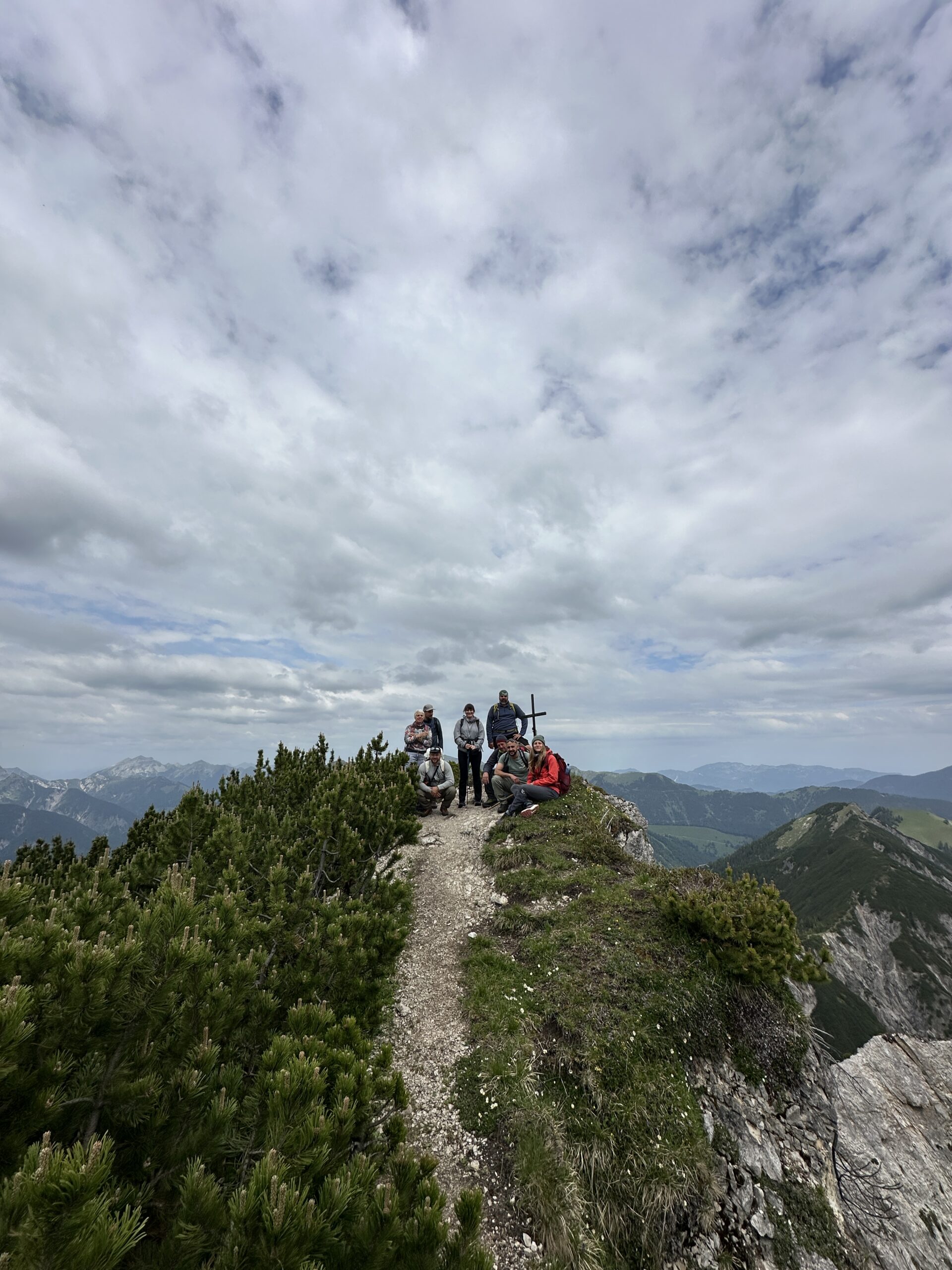 Eine Gruppe von sieben Personen sitzt und steht auf einem schmalen Berggrat neben einem Kreuz, vor dem sich ein bewölkter Himmel und ferne Berge abzeichnen, mit Blick auf die malerische Region Zaunreparaturen Ahornboden.