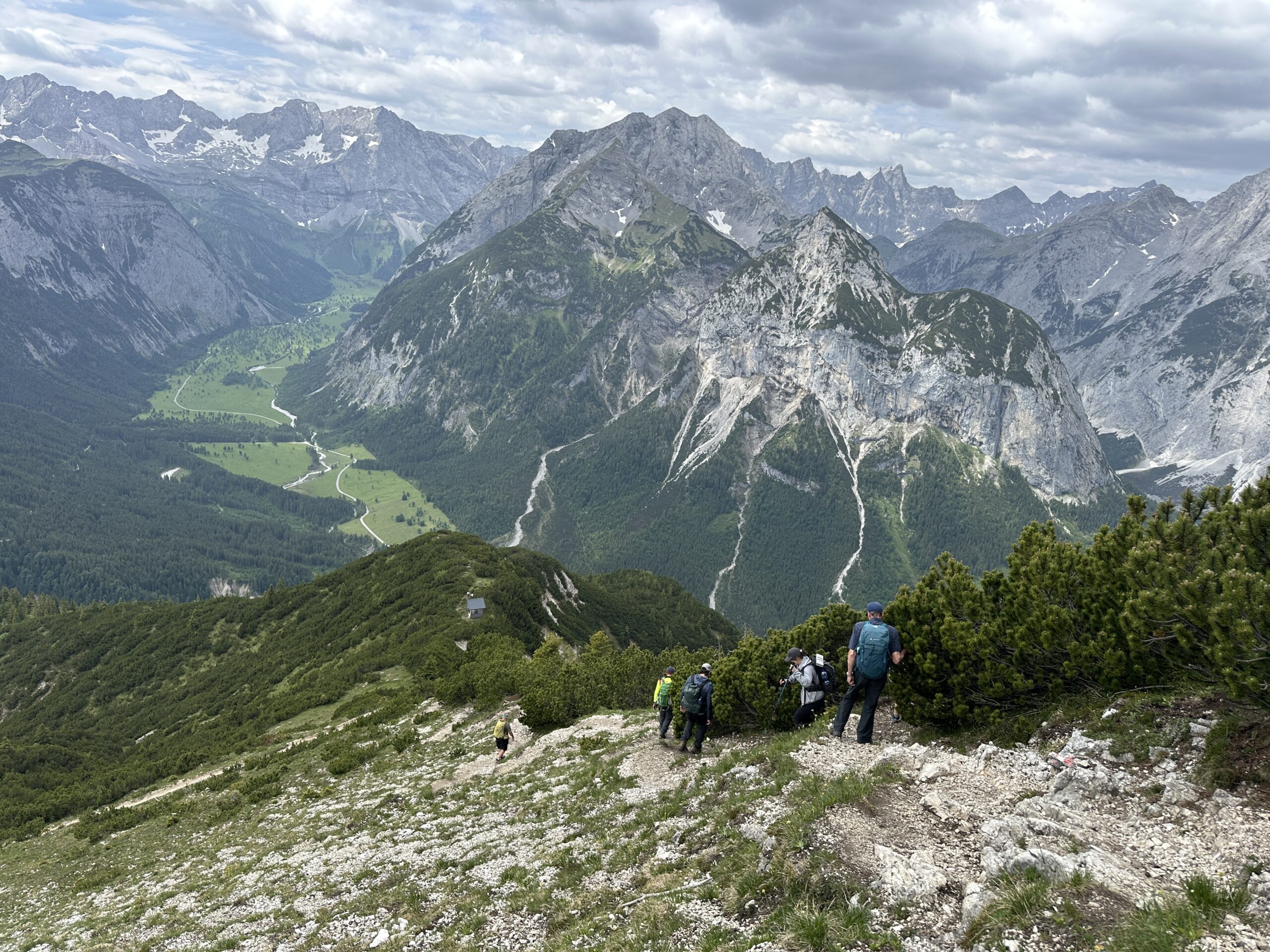Bergwaldprojekt Umweltbaustelle Almpflege Plumsjochhütte 2026 Team Karwendel Wanderer erklimmen einen felsigen, grünen Berghang in der Nähe von Zaunreparaturen Ahornboden, mit hohen, schroffen Gipfeln und einem Tal unter ihnen unter bewölktem Himmel.
