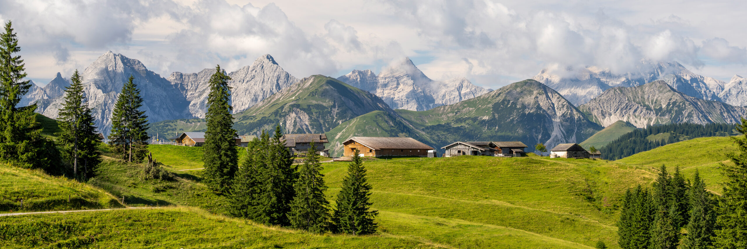 Eine kleine Ansammlung von Berghütten auf grünen Hügeln mit vereinzelten Bäumen, vor dem Hintergrund schroffer, schneebedeckter Berge und eines teilweise bewölkten Himmels.