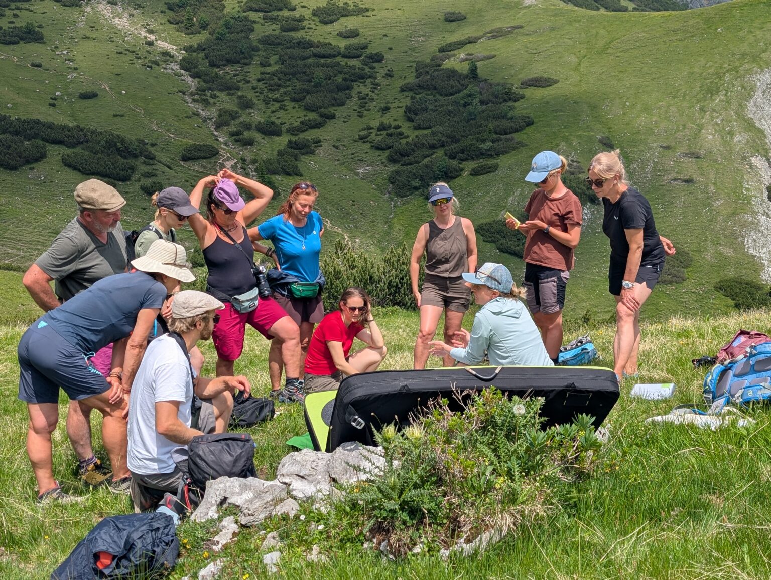 Eine Gruppe von Menschen hat sich im Freien auf einem grasbewachsenen Hang in der Karwendel-Region versammelt; einige stehen, andere sitzen und scheinen gemeinsam zuzuhören oder sich über etwas zu unterhalten. Rucksäcke und Outdoor-Ausrüstung sind zu sehen.