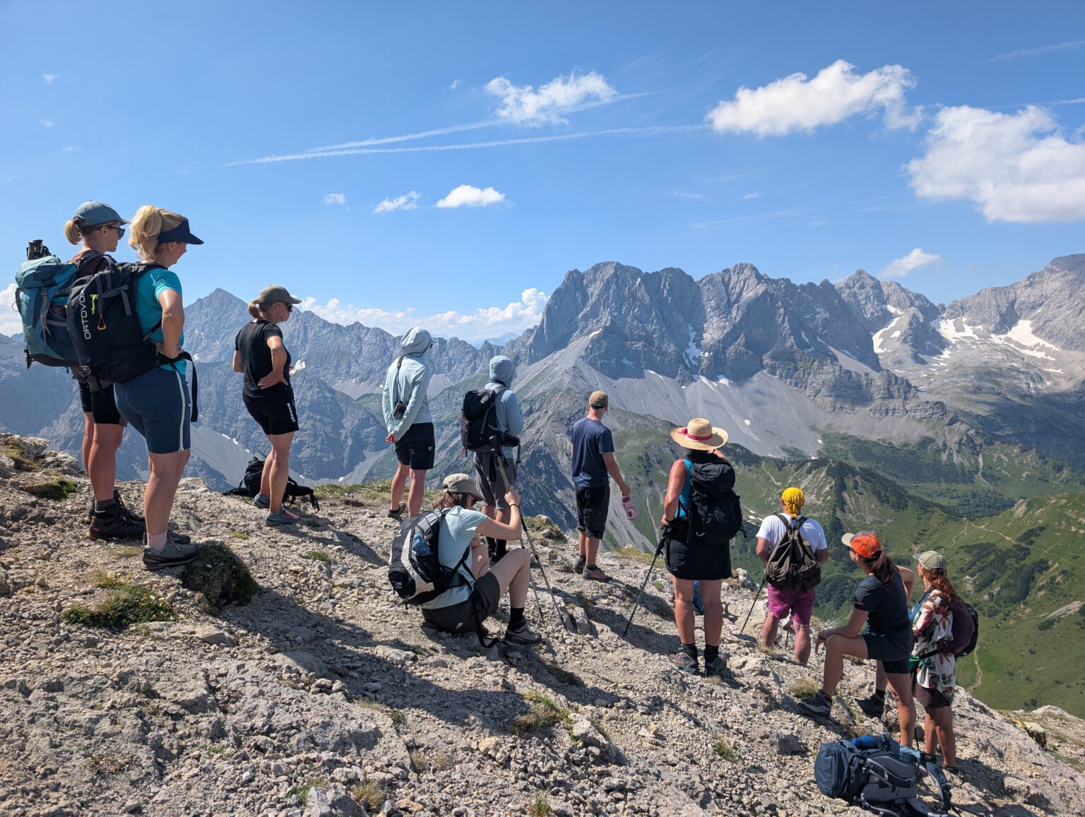 Eine Gruppe von Wanderern mit Rucksäcken und Wanderausrüstung steht und sitzt auf einem felsigen Berggrat im Karwendel und blickt unter strahlend blauem Himmel auf die fernen Gipfel.