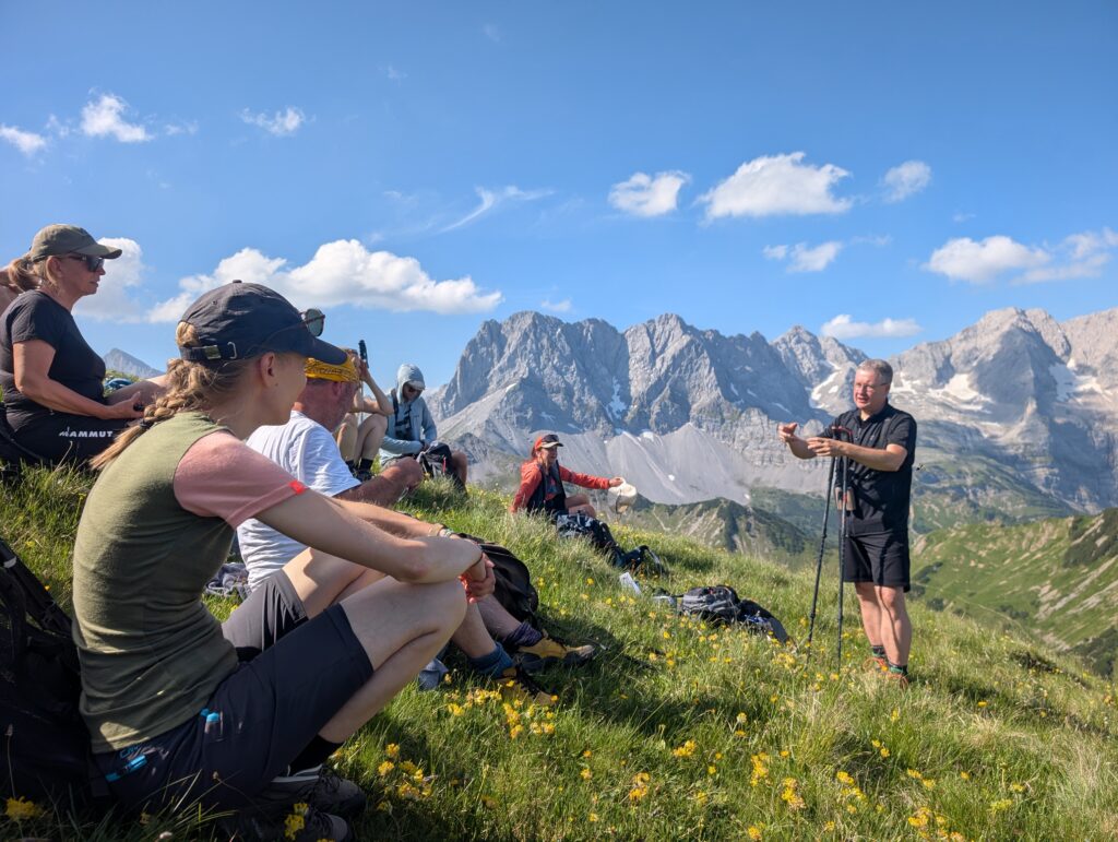 Eine Gruppe von Wanderern sitzt auf einem grasbewachsenen Hang im Karwendel und lauscht einem Mann, der dort steht und spricht; felsige Berge und ein blauer Himmel mit Wolken bilden die atemberaubende Kulisse.