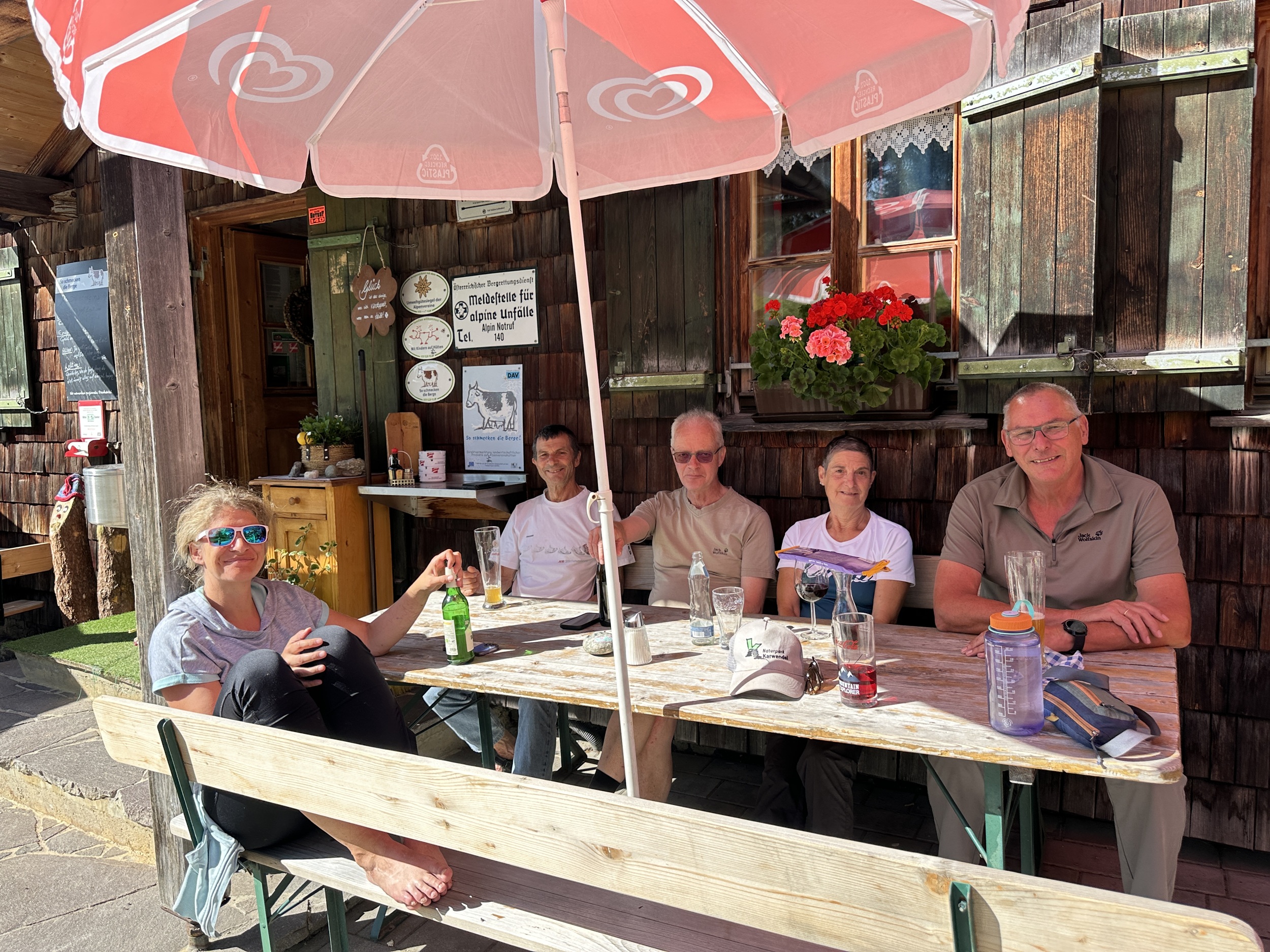 Bergwaldprojekt Umweltbaustelle Almpflege Plumsjochhütte 2026 Team Karwendel Fünf Personen entspannen sich an einem Holztisch im Freien unter einem roten Sonnenschirm, mit Getränken und Wanderausrüstung, vor dem rustikalen, mit Blumenkästen geschmückten Gebäude der Almpflege Martinsbergalm.