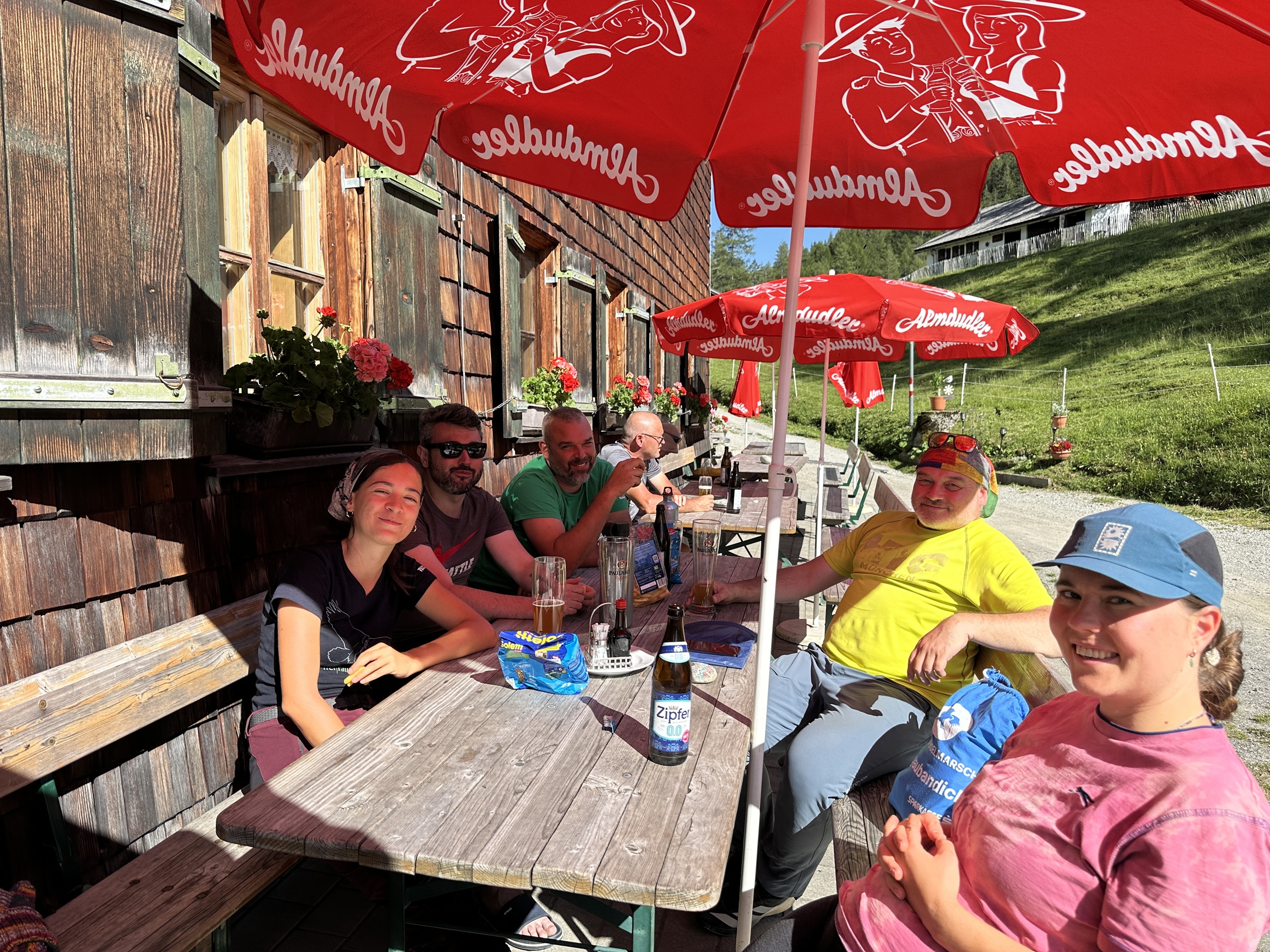 Bergwaldprojekt Umweltbaustelle Almpflege Plumsjochhütte 2026 Team Karwendel Eine Gruppe von sechs Personen sitzt unter roten Sonnenschirmen an einem Picknicktisch vor dem Holzgebäude der Almpflege Martinsbergalm und genießt an einem sonnigen Tag ein Getränk.