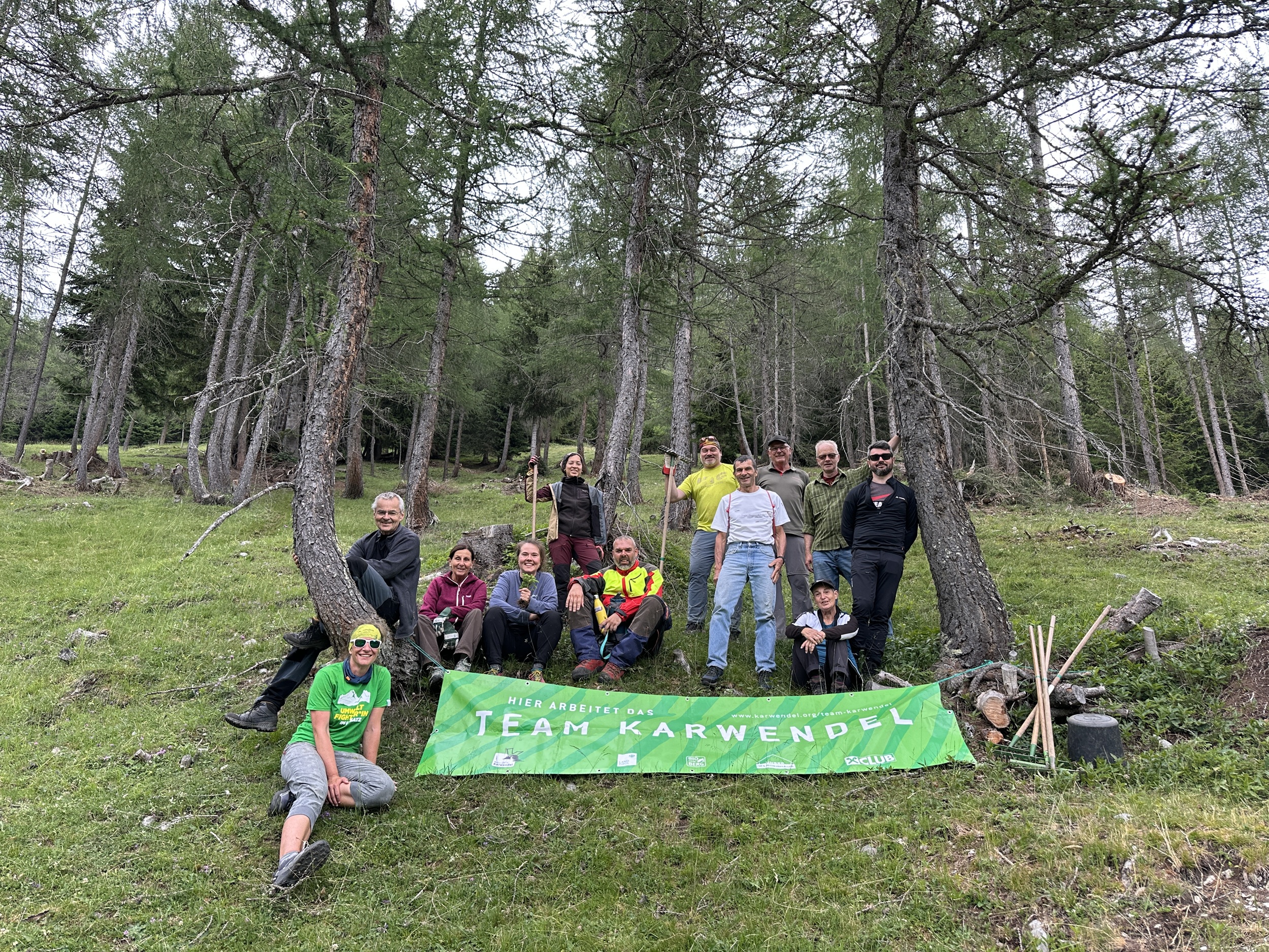 Eine Gruppe von Menschen posiert im Freien auf einer Waldlichtung in der Nähe der Plumsjochhütte; sie stehen und sitzen hinter einem grünen „Team Karwendel“-Banner. Vor ihnen liegen verschiedene Almpflege-Werkzeuge auf dem Gras.