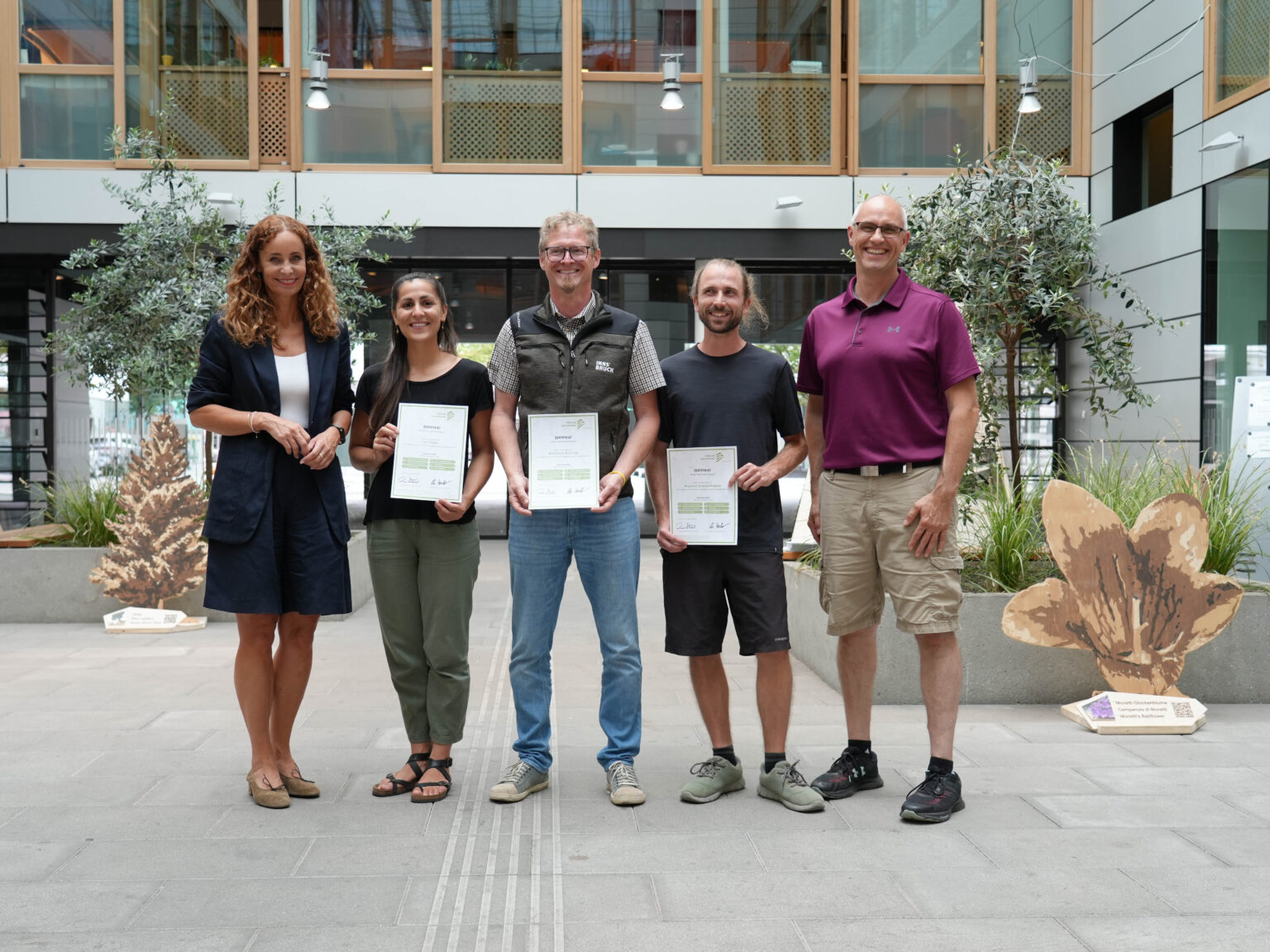 Fünf Erwachsene stehen in einem Raum, drei von ihnen halten Zertifikate in der Hand und posieren für ein Gruppenfoto in einem modernen Atrium mit Pflanzen und Tageslicht – die Szene fängt die Freude über den Erfolg nach ihrem Workshop zum Thema Karwendel ein.