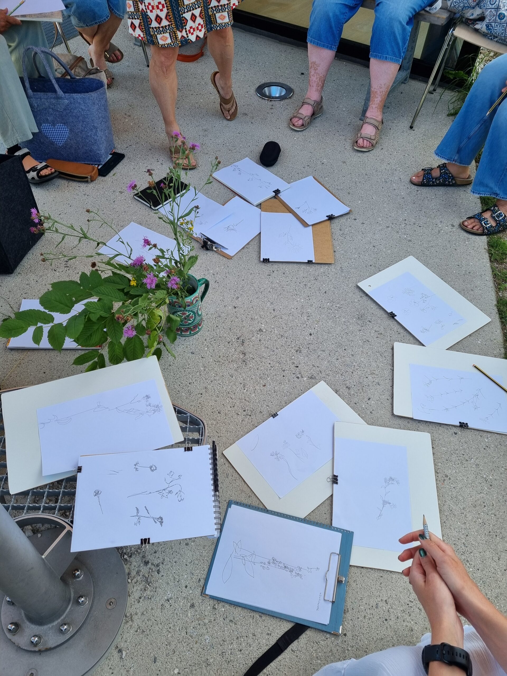 Die im Kreis sitzenden Menschen haben Skizzenbücher mit botanischen Zeichnungen auf den Boden gelegt und halten nachdenklich Details fest, die subtile Veränderungen im Zusammenhang mit dem Klimawandel im Gebirge offenbaren. In der Mitte steht eine Topfpflanze als Bezugspunkt. Zu sehen sind nur Beine und Hände.
