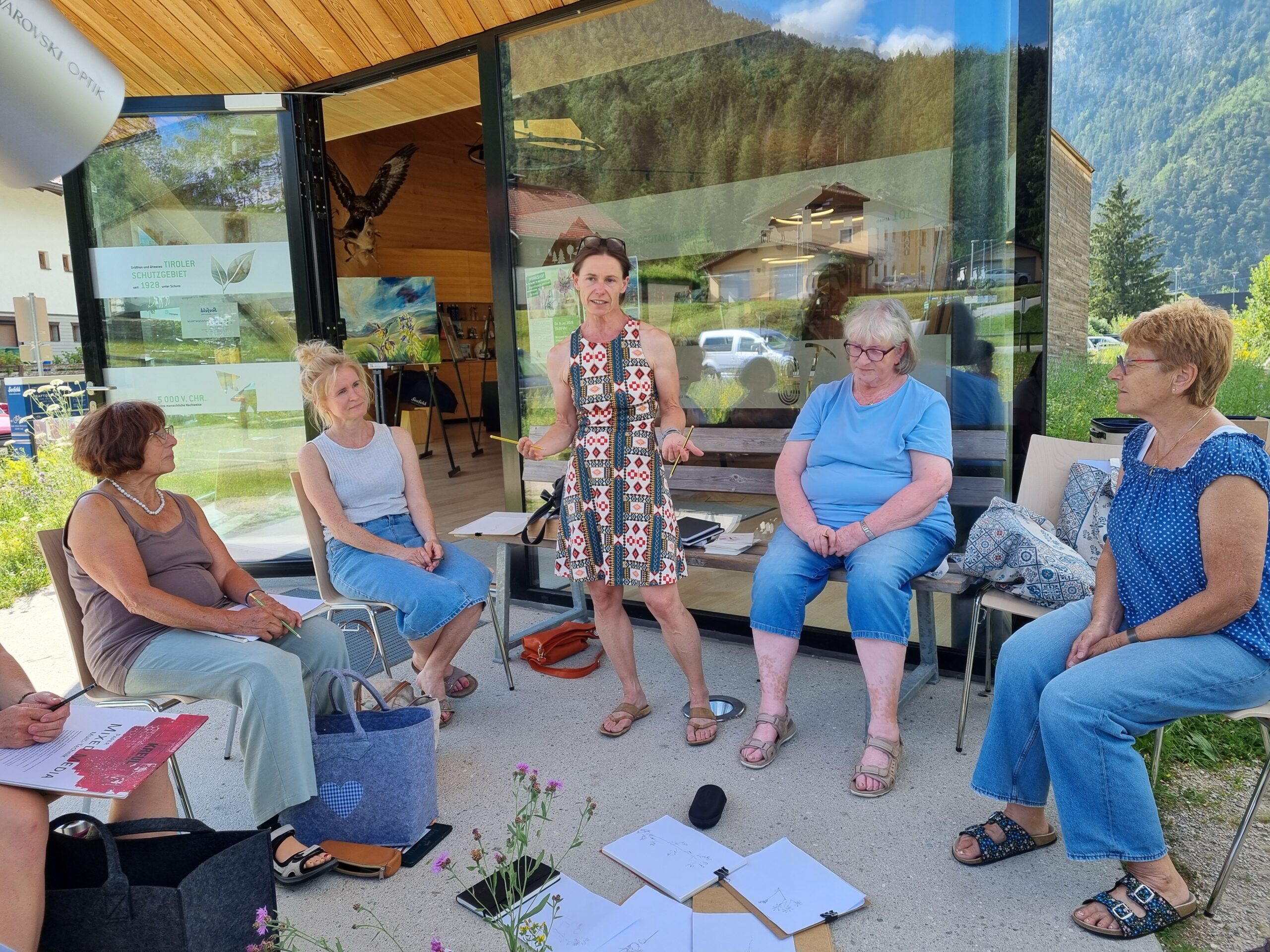 Fünf Frauen sitzen im Freien in einem Halbkreis in der Nähe eines Gebäudes mit Glaswänden und führen eine lebhafte Diskussion über den Klimawandel im Gebirge. In der Mitte der Gruppe liegen Papiere, ein Mikrofon und ein Tablet auf dem Boden, was darauf hindeutet, dass sie Forschungsergebnisse oder persönliche Erfahrungen im Zusammenhang mit dem Klimawandel in Bergregionen austauschen.