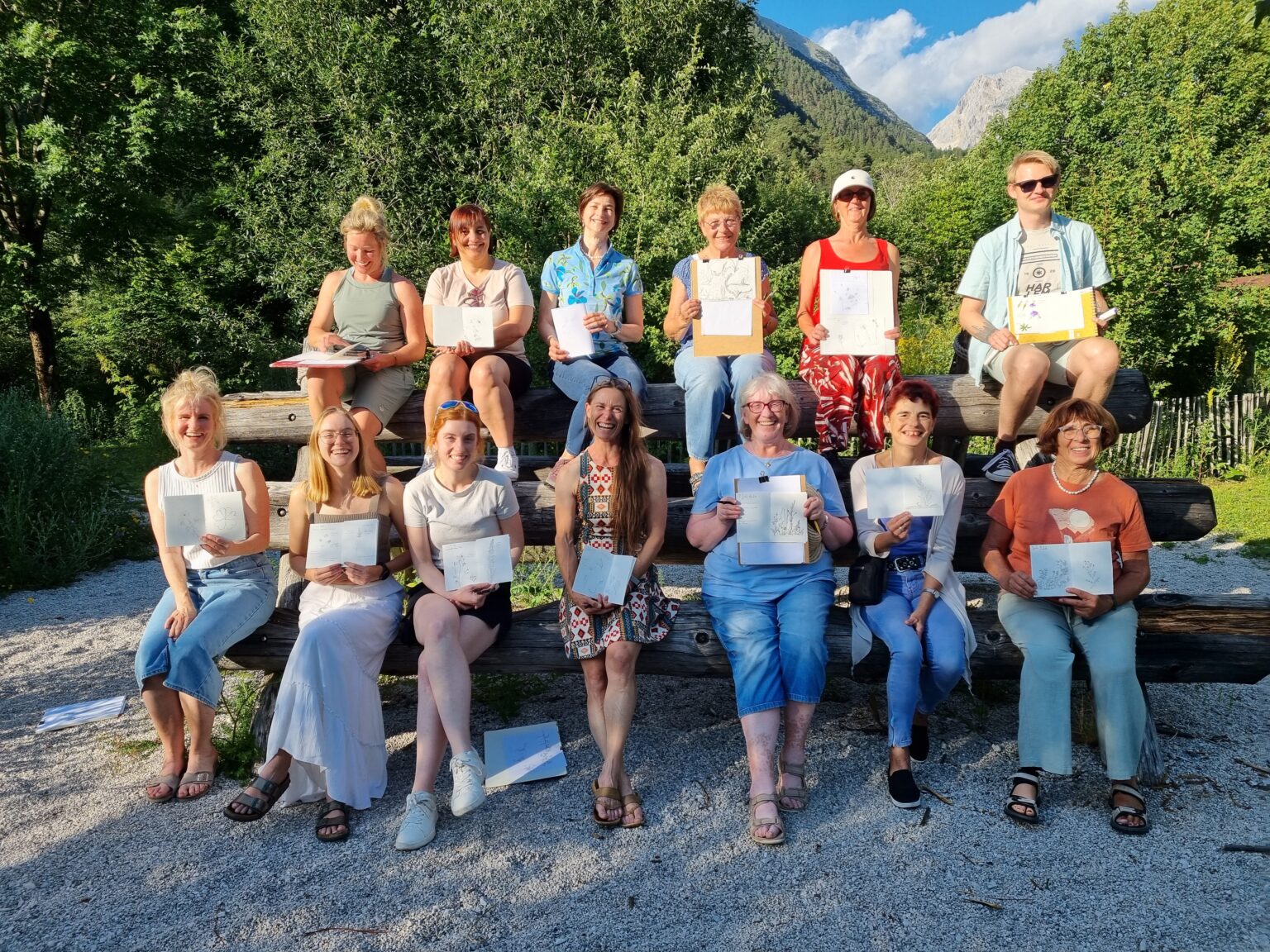 Eine Gruppe von vierzehn Personen posiert im Freien auf einer Holztreppe; jede hält eine Zeichnung oder ein Skizzenbuch zum Thema „Alpenblumen im Karwendel“ in der Hand, im Hintergrund sind Bäume und Berge zu sehen.
