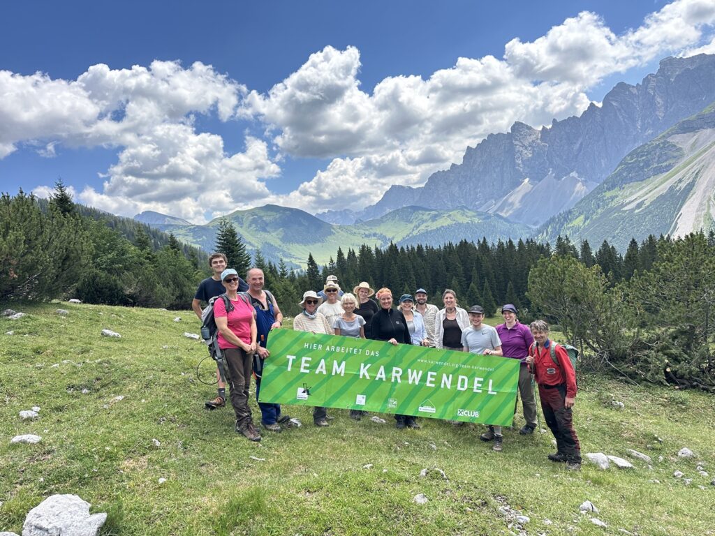 Eine Gruppe von Menschen im Freien hält ein grünes „Team Karwendel“-Transparent hoch, auf dem Berge, Bäume und ein bewölkter blauer Himmel im Hintergrund zu sehen sind, und feiert damit ihre Bemühungen im Rahmen der Almpflege Martinsbergalm.