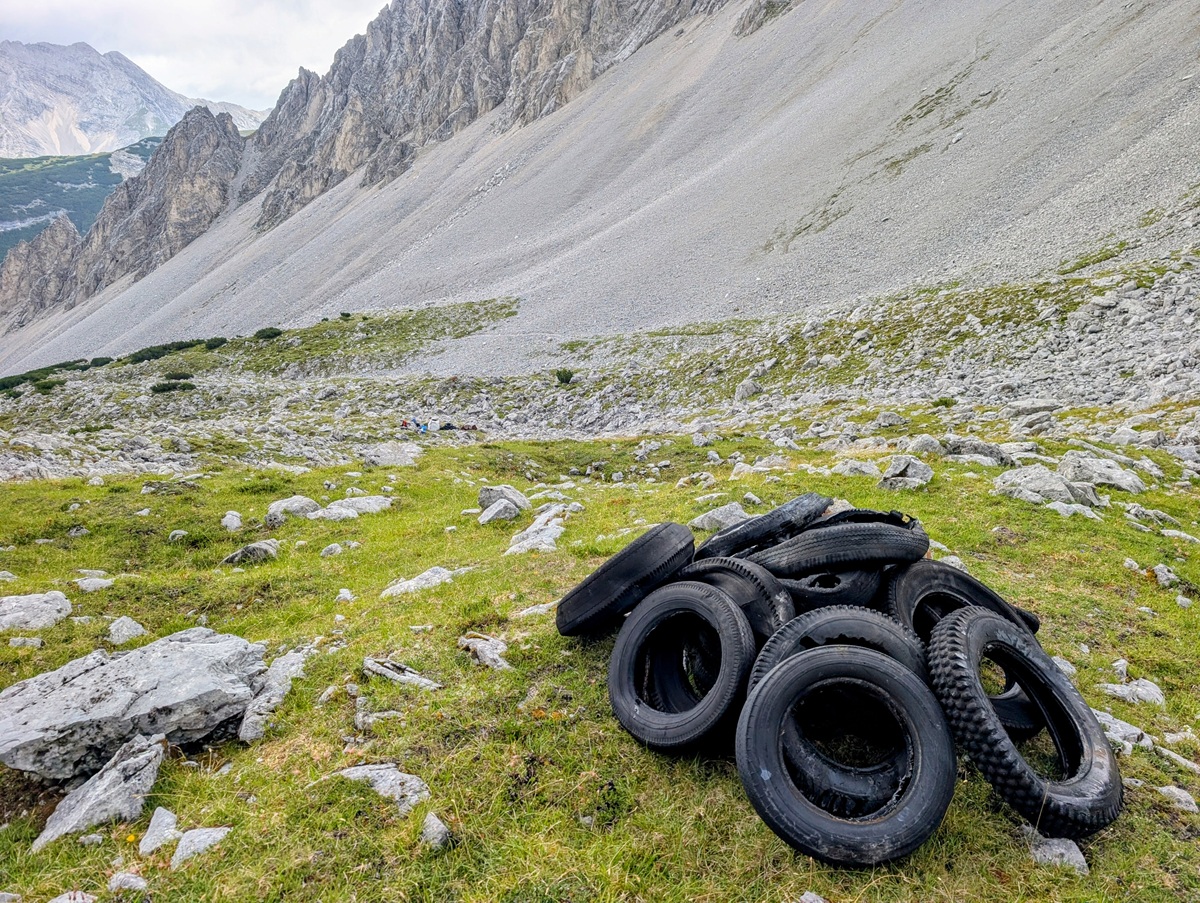 Unter einem bewölkten Himmel liegt ein Haufen ausrangierter Reifen auf felsigem, grasbewachsenem Gelände am Fuße eines steilen Berghangs im Karwendel.