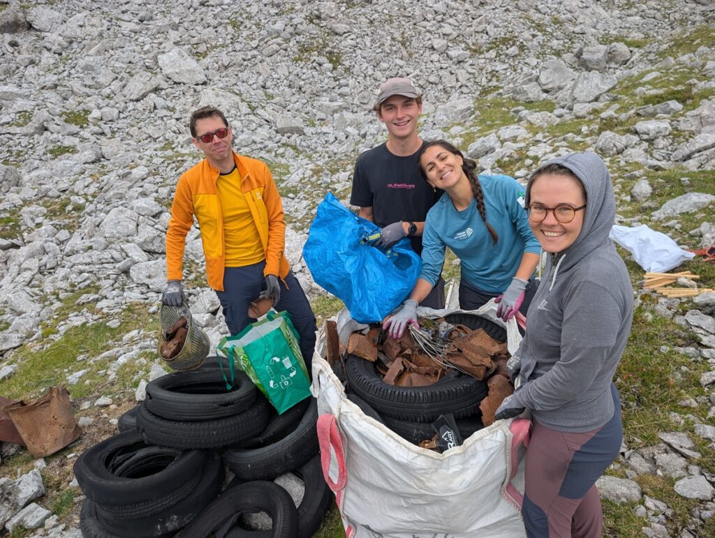 Vier Personen sammeln im Karwendel im Freien Müll – darunter alte Reifen und Metallschrott – und packen ihn inmitten der felsigen Landschaft in große Säcke.