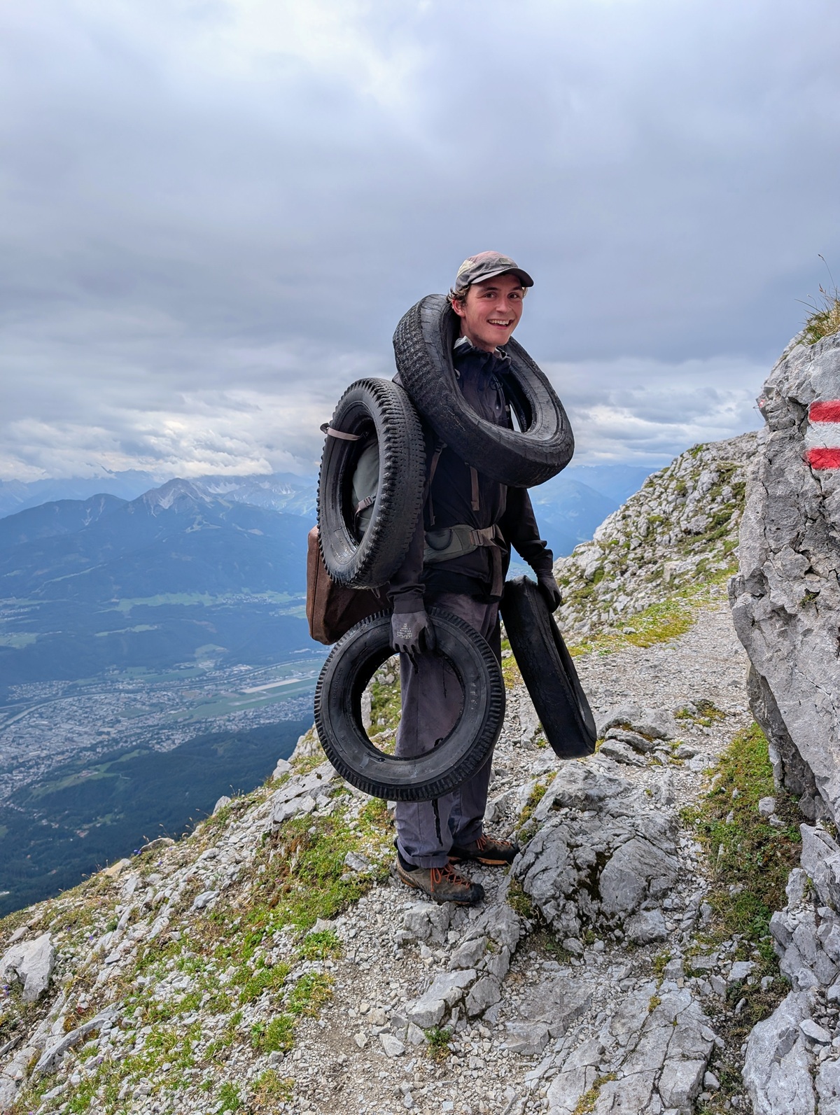 Eine Person steht auf einem felsigen Bergpfad im Karwendel, trägt mehrere Reifen und hält einen weiteren in der Hand; der Himmel ist bewölkt, und im Tal darunter ist eine Stadt zu sehen.