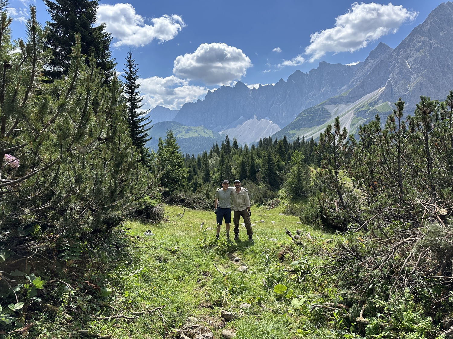 Zwei Menschen stehen auf einer von Kiefern umgebenen Wiese auf der Hochalm, im Hintergrund sind Berge und ein teilweise bewölkter Himmel zu sehen – eine friedliche Szene, die an traditionelle Almpflegepraktiken erinnert.