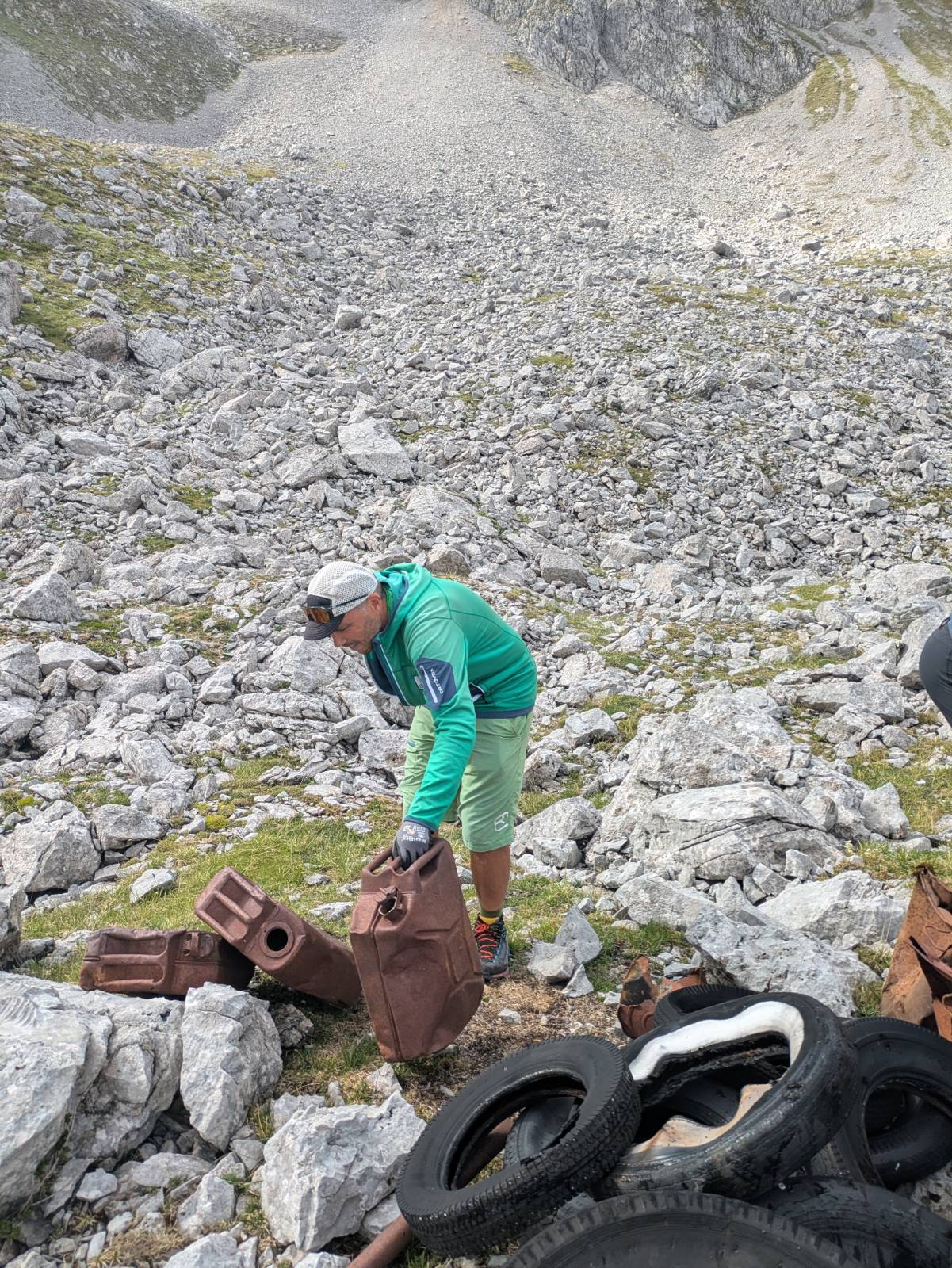 Eine Person in Outdoor-Kleidung hantiert an einem felsigen Hang im Karwendel mit verrosteten Benzinkanistern und alten Reifen, umgeben von Steinen und spärlichem Gras.