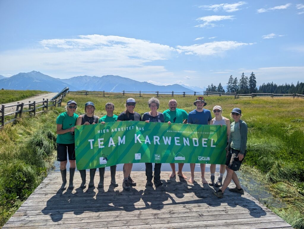 Eine Gruppe von zehn Personen steht im Freien auf einem Holzsteg und hält ein grünes Transparent mit der Aufschrift „Team Karwendel“ in den Händen; im Hintergrund sind Berge und Wiesen zu sehen.