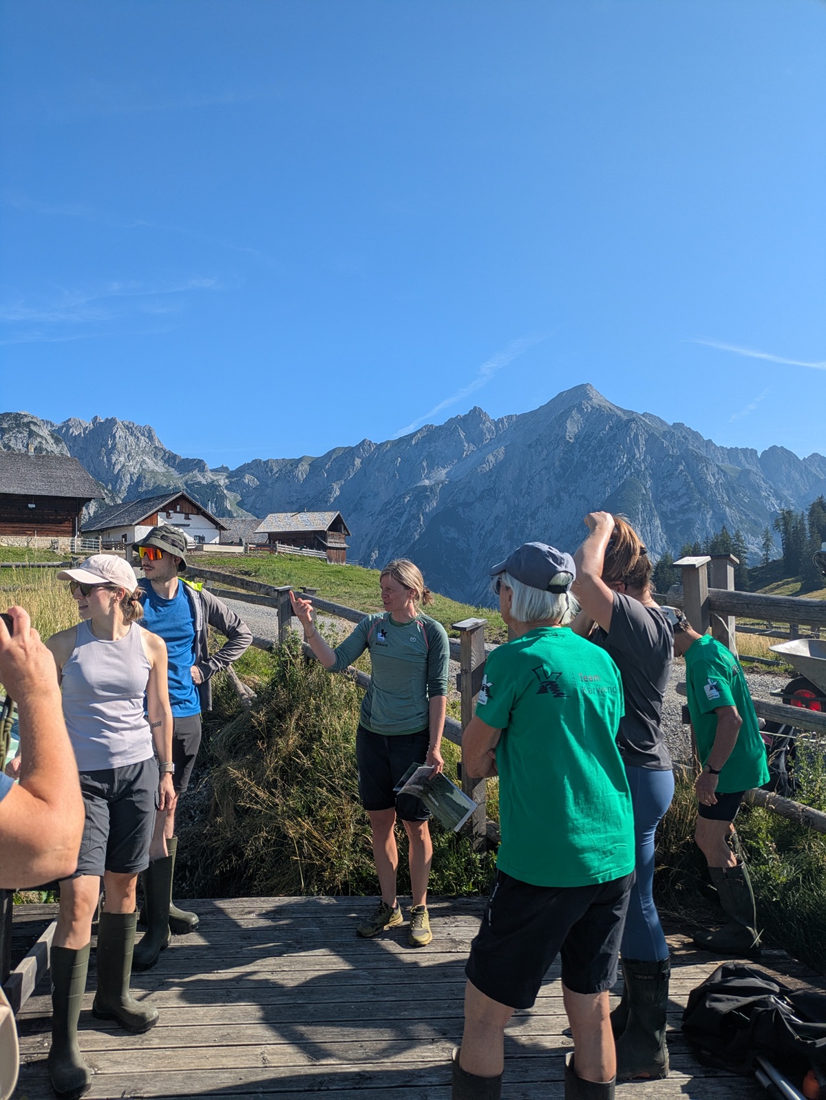 Eine Gruppe von Menschen in Outdoor-Kleidung steht auf einer Holzterrasse in einer bergigen Gegend unter einem strahlend blauen Himmel.