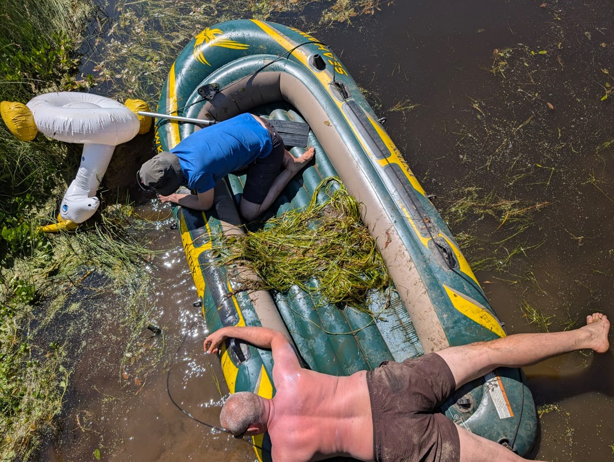 Zwei Menschen ruhen sich auf und neben einem Schlauchboot in seichtem, mit Wasserpflanzen bewachsenem Wasser aus; einer liegt mit dem Gesicht nach unten auf dem Schlauchboot, der andere ist bis zur Hälfte im Wasser versunken und hat einen Sonnenbrand. In der Nähe schwimmt ein aufblasbarer Schwan.