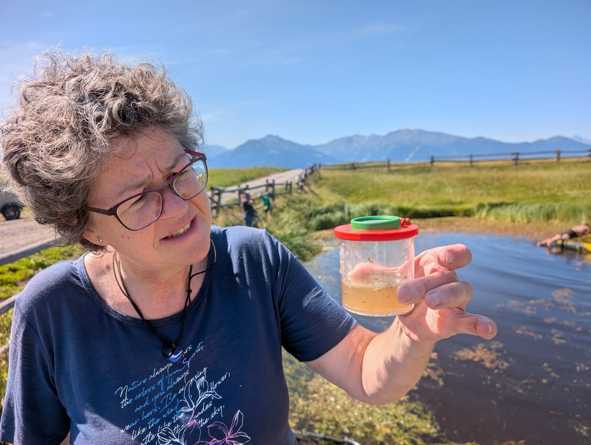 Eine Frau mit grauen Locken betrachtet einen durchsichtigen Behälter mit Wasser; sie steht im Freien in der Nähe eines Teiches, im Hintergrund sind Berge und ein blauer Himmel zu sehen.