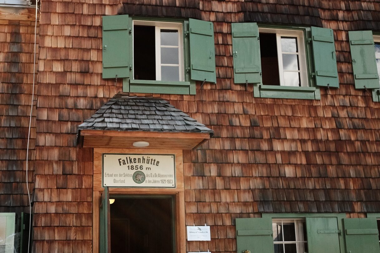Bergwaldprojekt Umweltbaustelle Almpflege Plumsjochhütte 2026 Team Karwendel Eine Gebäudefassade aus Holz mit grünen Fensterläden, offenen Fenstern und einem Schild mit der Aufschrift „Falkenhütte 1856 m“ über dem Eingang – typisch für die traditionelle Architektur der Anger-Hochalm im Stil der Almpflege. Im Vordergrund ist ein Teil eines Kopfes zu sehen.