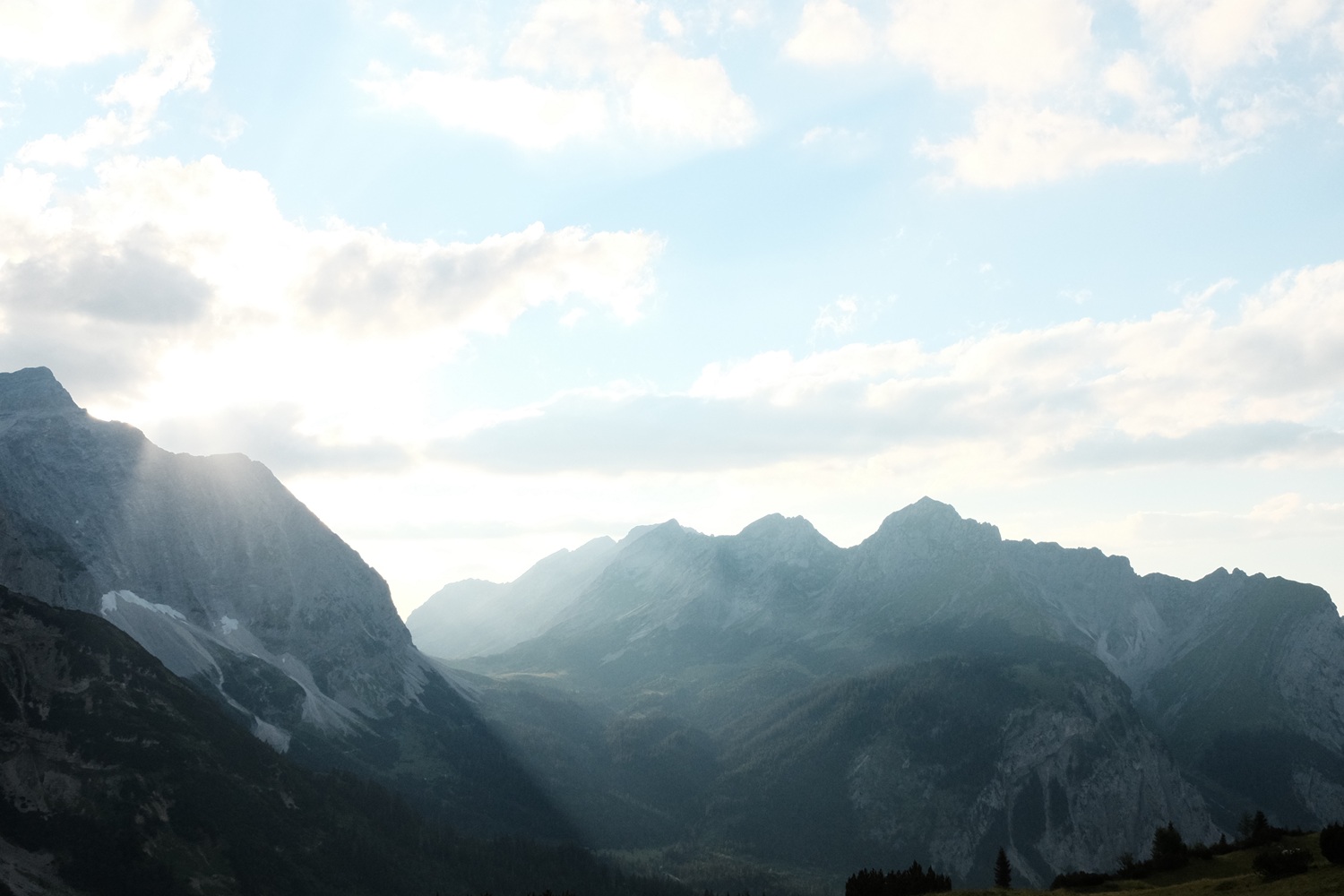Bergwaldprojekt Umweltbaustelle Almpflege Plumsjochhütte 2026 Team Karwendel Eine Bergkette, durch deren Wolken Sonnenstrahlen scheinen und die felsigen Gipfel und bewaldeten Täler unter einem strahlenden Himmel erhellen – das erinnert an die beschauliche Almpflege Anger Hochalm, wo Almwiesen und imposante Gipfel in harmonischer natürlicher Schönheit aufeinander treffen.
