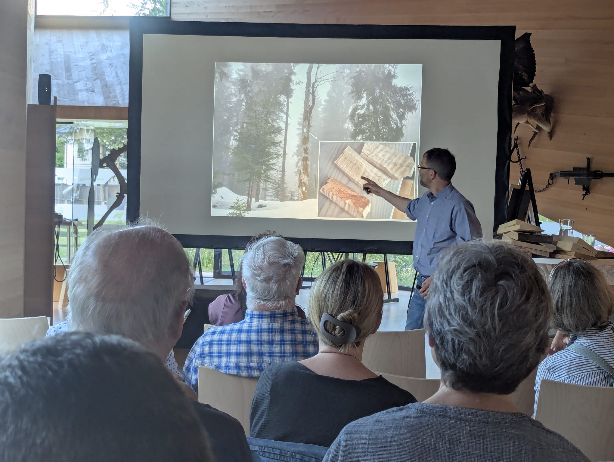 Ein Mann steht vor einem sitzenden Publikum, hält einen Vortrag über Holz und Wälder und zeigt dabei auf Bilder zum Thema Altlasten, die auf einer Leinwand projiziert werden.