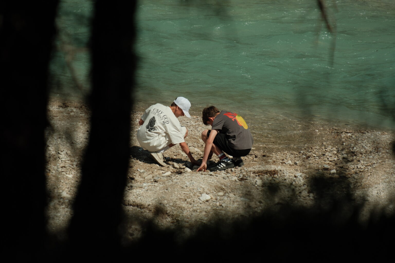 Zwei Menschen hocken an einem felsigen Ufer am türkisfarbenen Wasser und beschäftigen sich mit Umweltbildung, während sie die Felsen untersuchen oder durchsuchen. Bäume verdecken teilweise den Vordergrund.