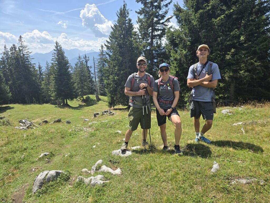 Drei Menschen stehen auf einem grasbewachsenen, felsigen Hang im Herzen des Karwendels, in Wanderkleidung und umgeben von Kiefern und majestätischen Bergen unter einem teilweise bewölkten Himmel.