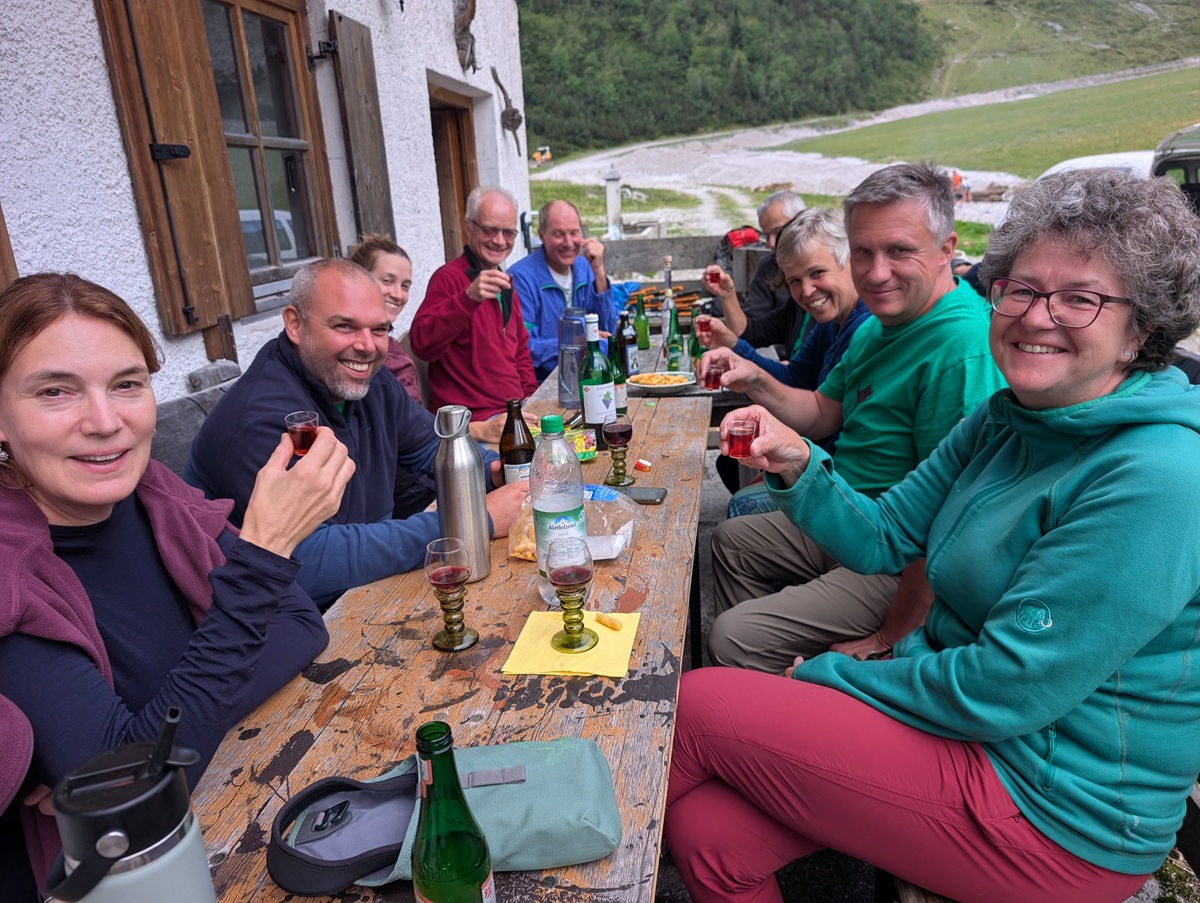Eine Gruppe von Menschen sitzt an einem rustikalen Tisch im Freien auf der Lalidersalm, lächelt und erhebt die Gläser zum Anstoßen. Auf dem Tisch stehen Flaschen und Becher, im Hintergrund ist eine grasbewachsene Landschaft zu sehen.
